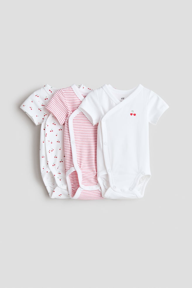 Three short-sleeved, wrapover bodysuits are displayed, made from soft cotton jersey and featuring press-studs down one side and at the crotch. The set includes a white bodysuit with a cherry graphic, a white bodysuit with red stripes, and a white bodysuit with a red cherry pattern. - 1