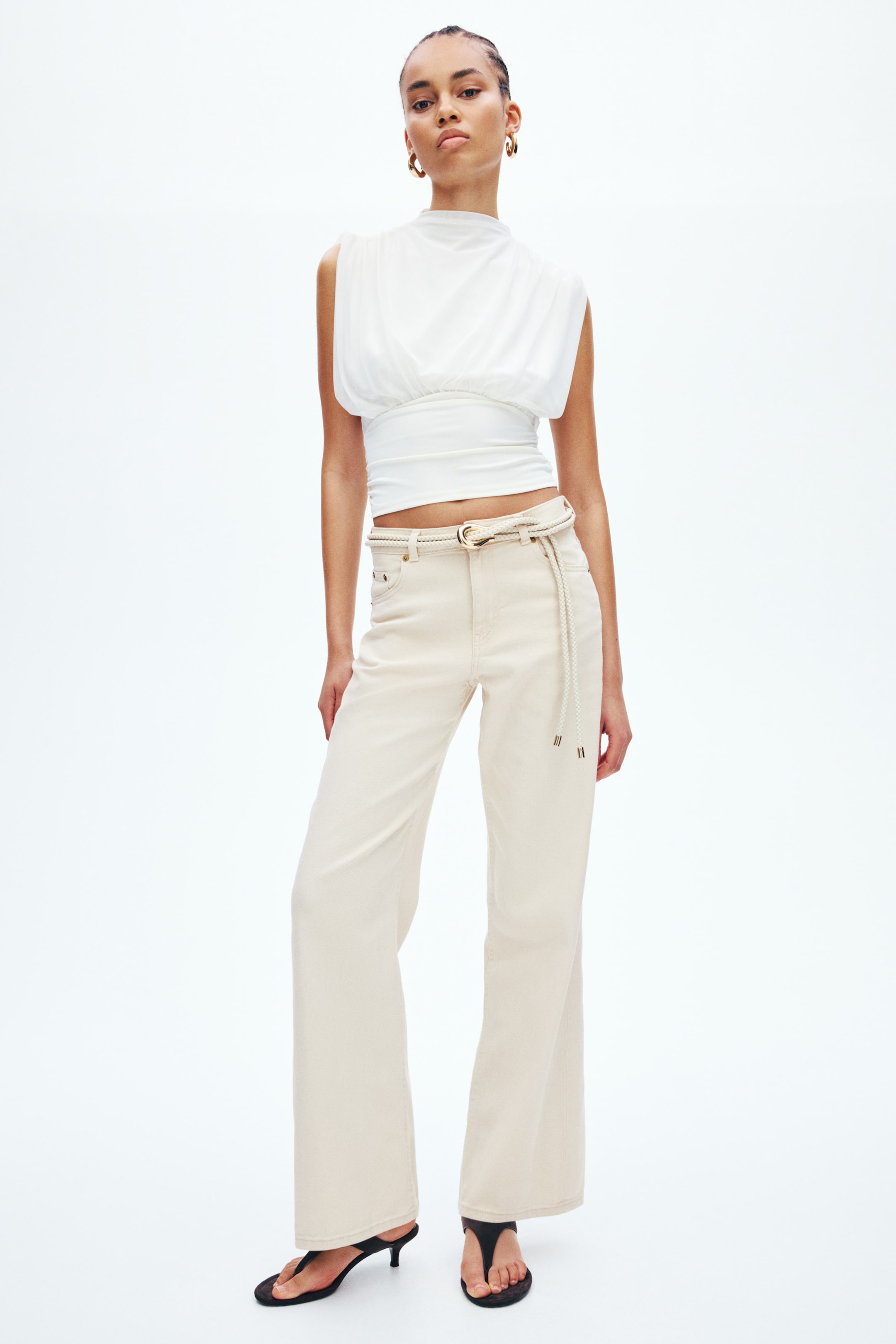 View larger image: Light beige wide-leg jeans feature five-pocket styling and visible belt loops. A braided rope belt with a gold-toned buckle threads through the loops, hanging loosely. The full-length hems fall to the floor.
