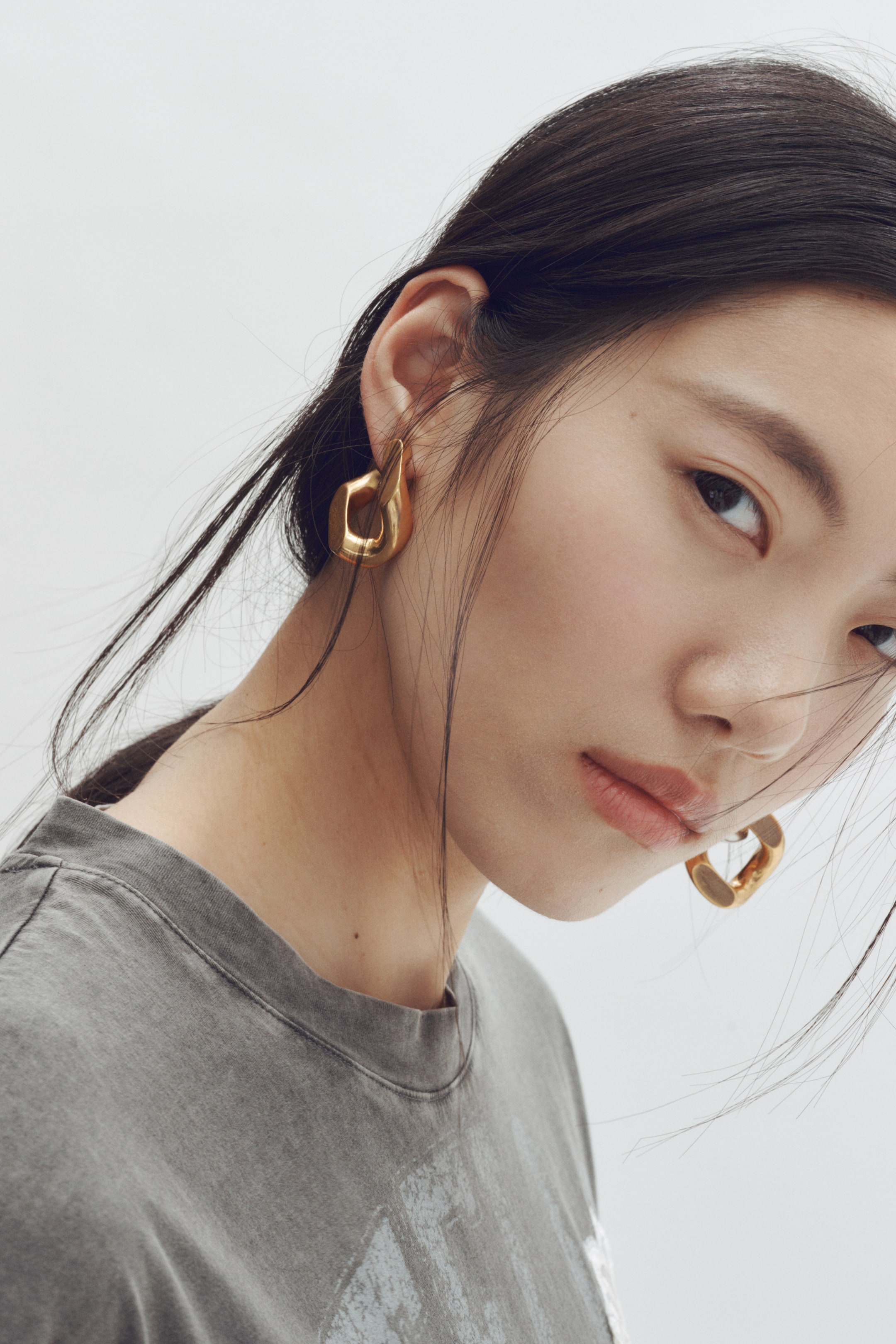 View larger image: Gold-toned sculptural earrings are being worn, featuring an abstract, curved design with a smooth, polished finish. One earring is resting on the earlobe from a side-front angle, while the other is showcasing its hollowed-out, rounded shape from a side view.