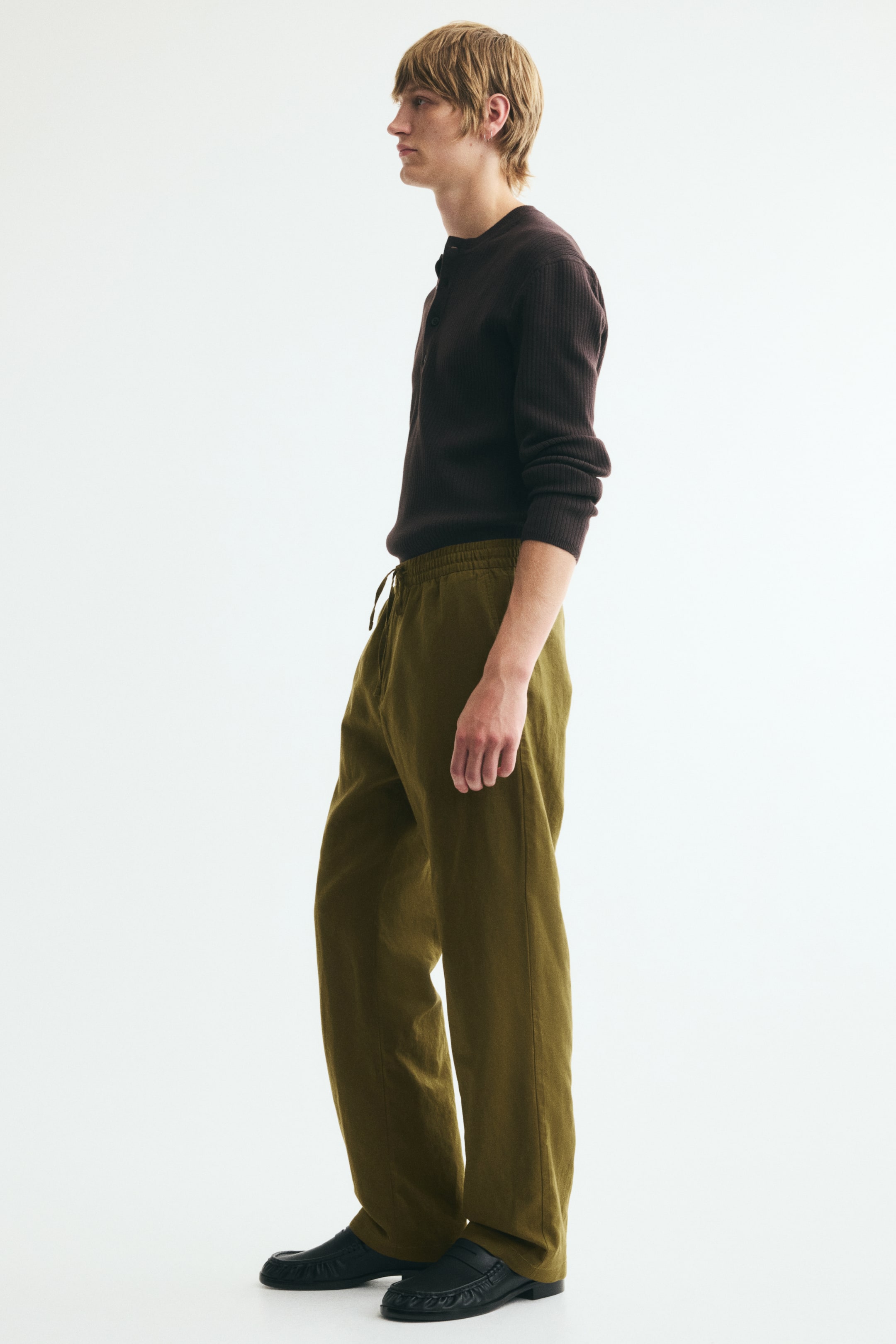 View larger image: Olive green cotton and linen trousers, featuring a visible drawstring at the covered elasticated waist, are worn by the model. The trousers show a regular fit with a straight-leg silhouette, highlighting the relaxed drape of the fabric, while the model stands and pairs them with a dark long-sleeved shirt and black slip-on shoes.