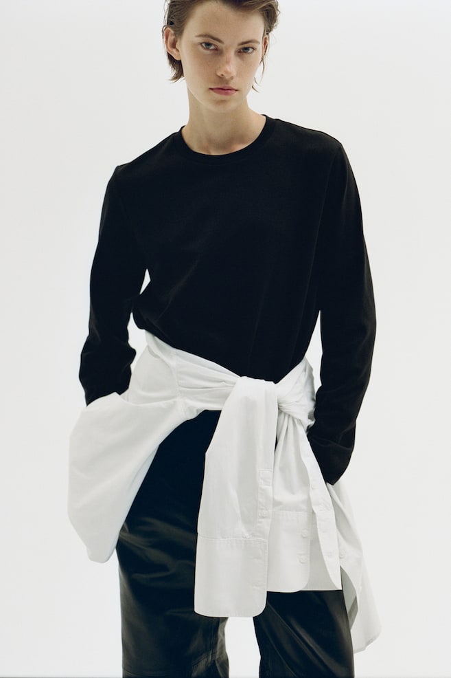 Een zwart topje met lange mouwen en een ronde halslijn wordt van voren getoond, waarbij de ontspannen en rechte pasvorm zichtbaar is. Het topje is gestyled met een wit overhemd met knoopsluiting dat om de taille is geknoopt, over een donkere broek. - 1