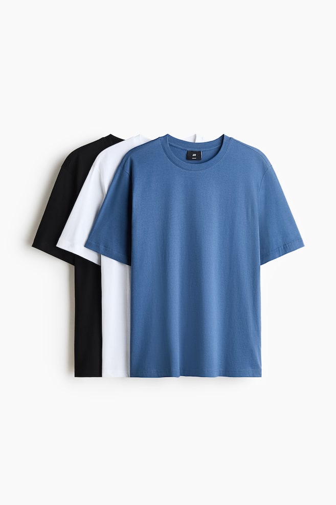 Three regular-fit cotton jersey T-shirts in blue, white and black are displayed hanging in a layered, front-facing arrangement. The blue T-shirt in the foreground showcases a round, rib-trimmed neckline, short sleeves and a straight hem, while the white and black T-shirts are layered behind it. - 1