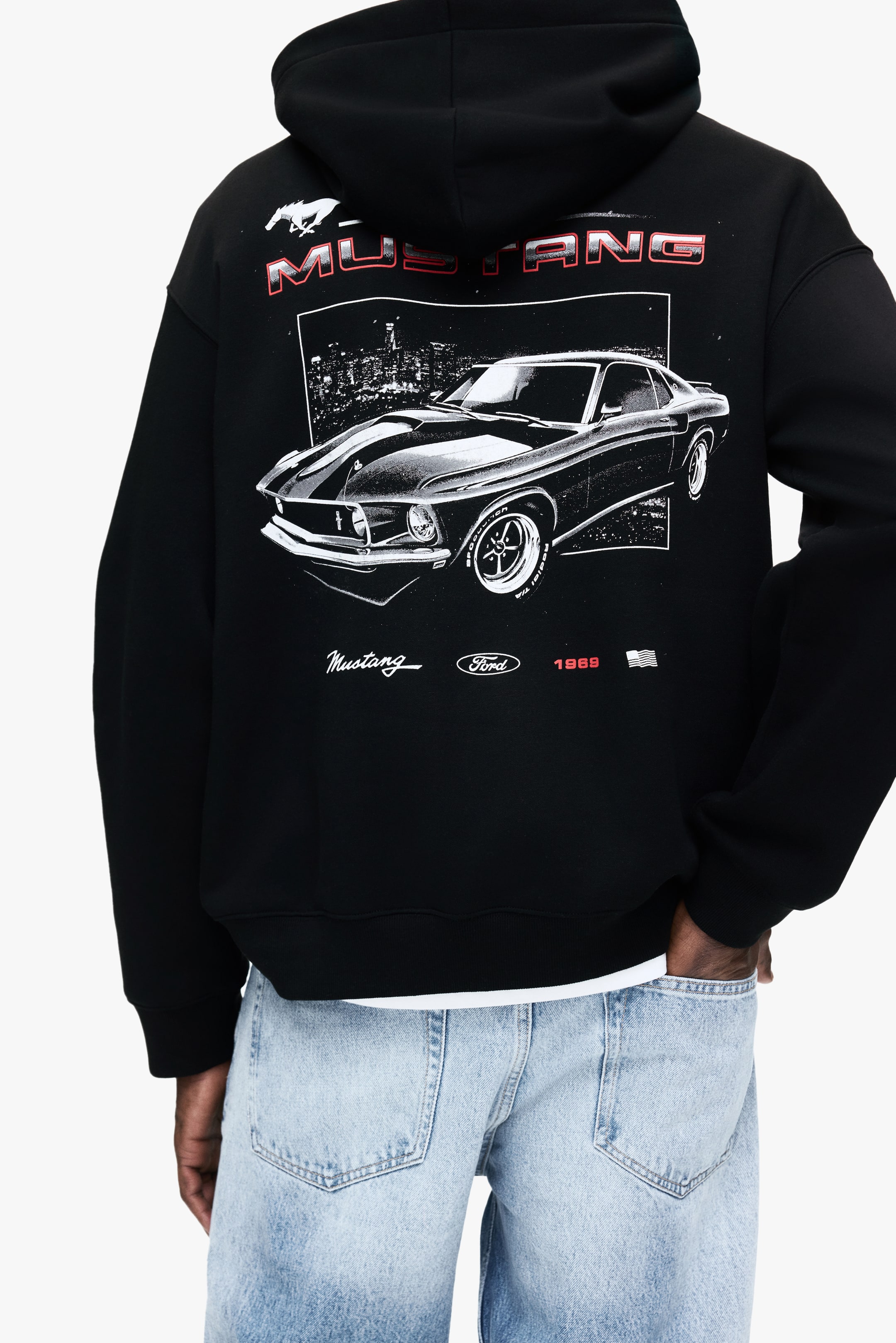 Grösseres Bild ansehen: Ein schwarzes Kapuzen-Sweatshirt mit einem grossen Grafikdruck eines klassischen Ford Mustang Autos, dem Text "MUSTANG", einem Pferdelogo, "Ford", "1969" und einem amerikanischen Flaggen-Icon auf der Rückseite wird vom Modell getragen, das eine Hand in die Tasche der hell gewaschenen Jeans steckt, die darunter getragen wird.