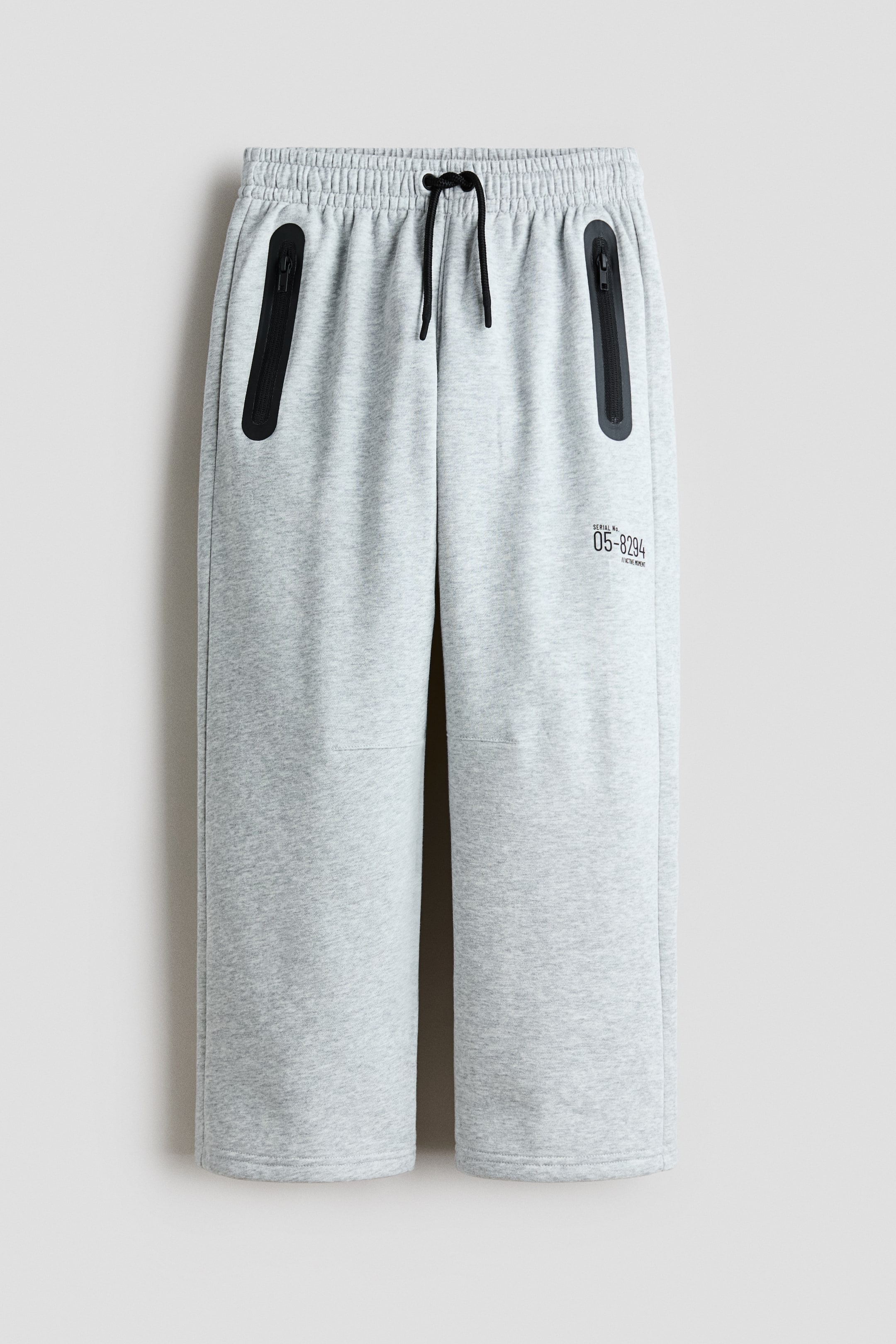 Agrandir l'image: Shorts en molleton gris chiné clair orientés vers l'avant, avec une taille élastiquée et un cordon de serrage noir. Deux poches zippées noires inclinées sont visibles sur le devant supérieur, et le petit texte noir "SERIAL No. 05-8294" et "ACTIVE WEAR" est imprimé sur la cuisse droite.