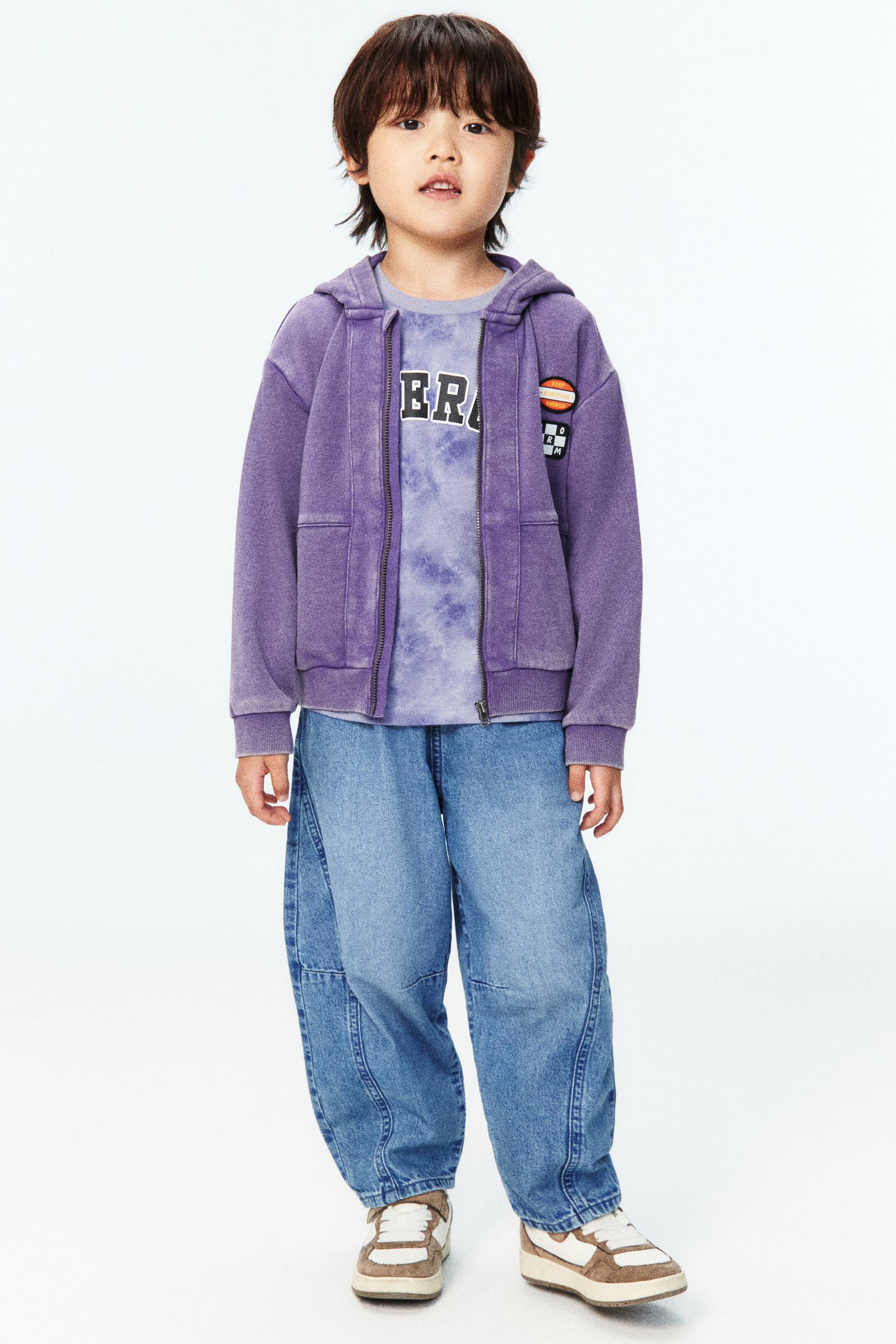 Xem ảnh lớn hơn: Một chiếc áo hoodie có khóa kéo màu tím nhạt, có khóa kéo phía trước, mũ trùm đầu, cổ tay áo bo gân và các miếng dán trên ngực, đang được người mẫu mặc. Nó được kết hợp với một chiếc áo thun tie-dye màu tím nhạt có chữ "ERA", và được phối với quần jeans ống rộng màu sáng và giày thể thao cổ thấp màu nâu và trắng.