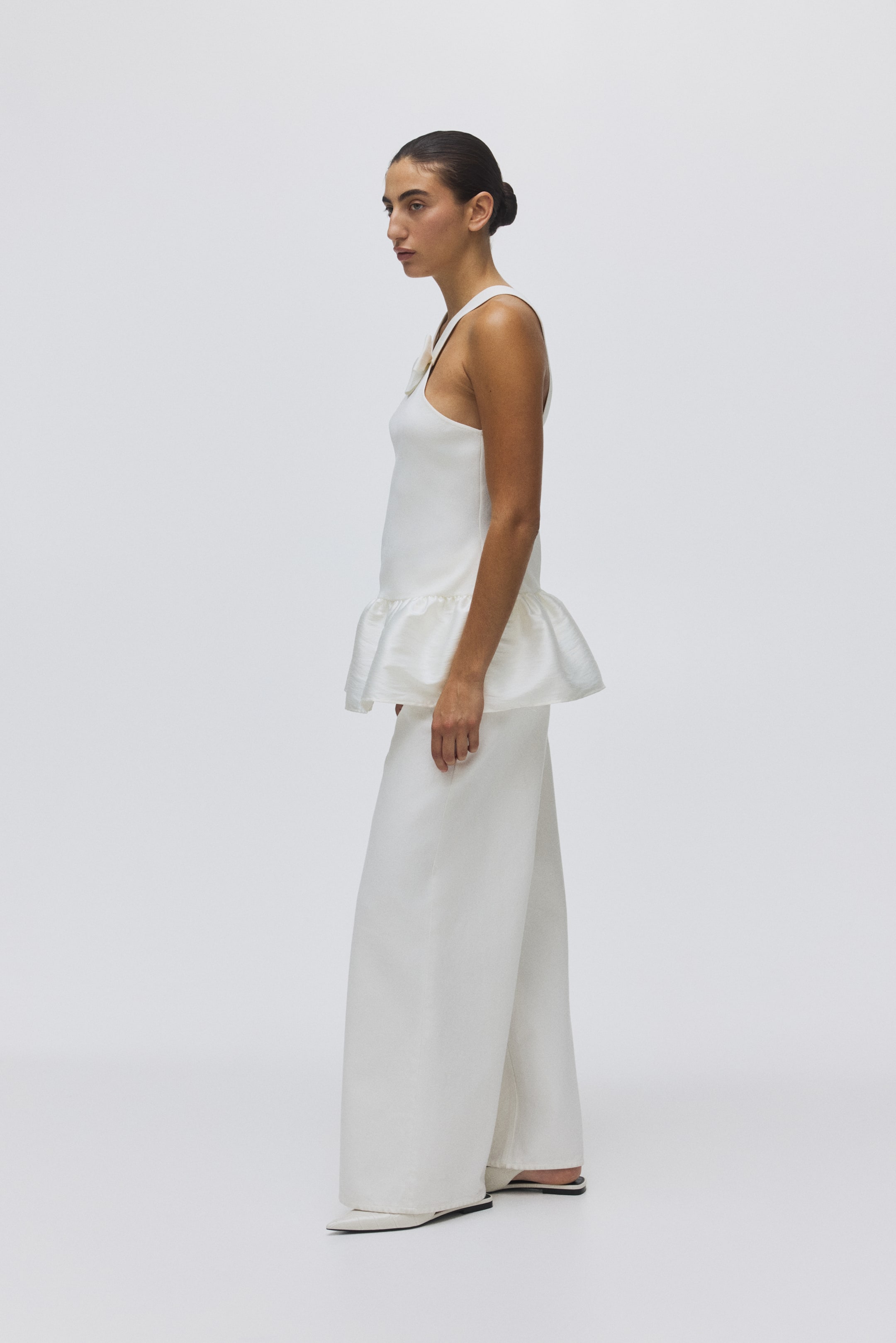 View larger image: An all-white ensemble is being shown from a side profile. A wide-strap sleeveless top with a gathered peplum hem is appearing over wide-leg trousers, while white pointed-toe flats are completing the look.