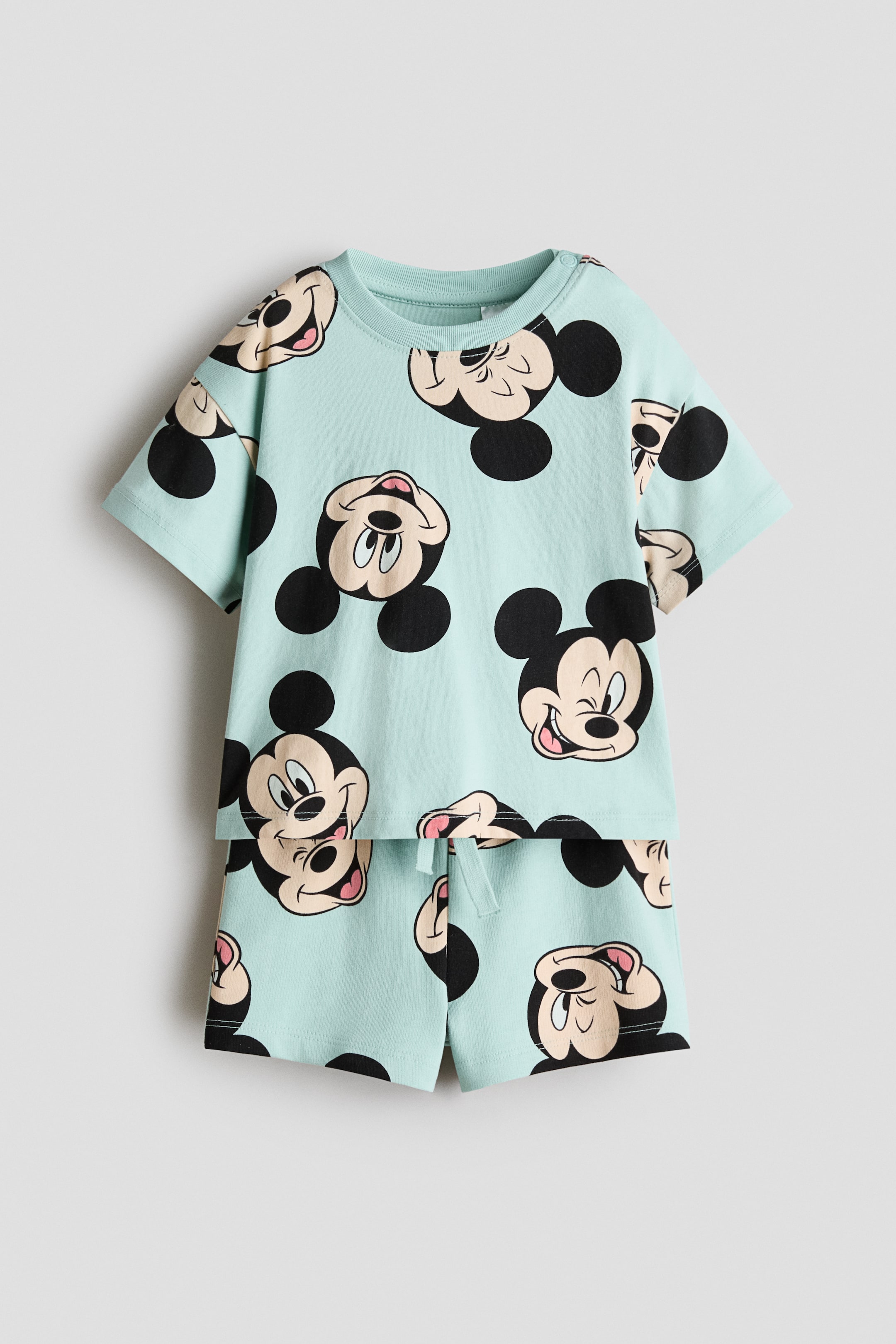 View larger image: A 2-piece printed set, facing front, features a light blue short-sleeved t-shirt and matching shorts, both adorned with an all-over print of Mickey Mouse faces in various expressions and orientations. The t-shirt is crafted with a ribbed crew neck and snap buttons on the left shoulder, while the shorts boast an elasticated waistband with a white drawstring.