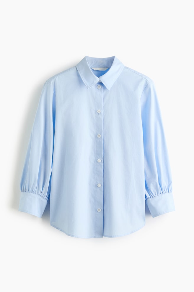 A light blue shirt is presented front-facing, featuring a pointed collar and a button-front placket with white buttons. It is designed with long sleeves that are ending in gathered cuffs and a curved hemline. - 1