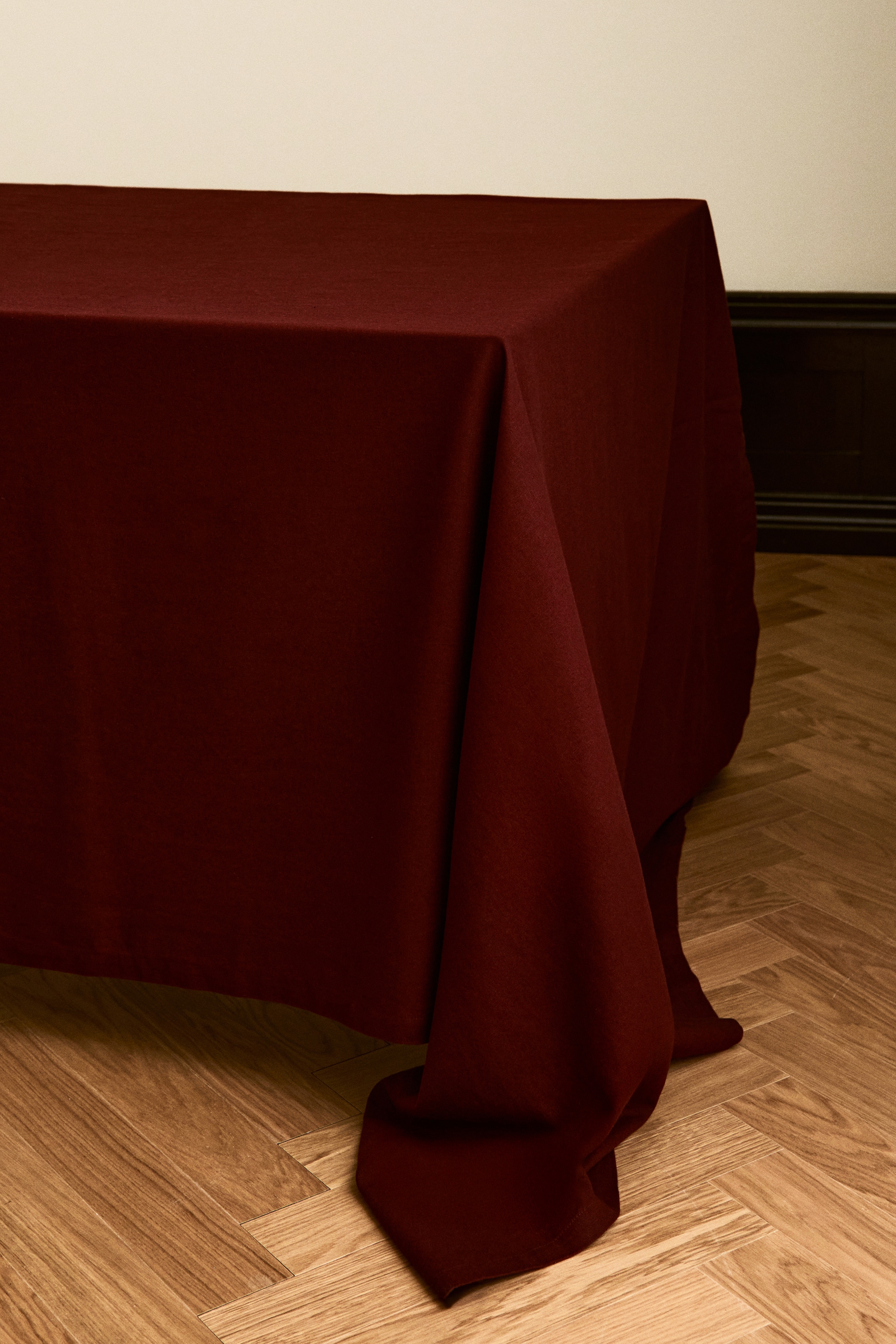 A woven tablecloth in a cotton and linen blend is shown folded from the front in a deep burgundy hue. The fabric is showcasing a textured matte finish and is featuring a visible folded hem along the edges.