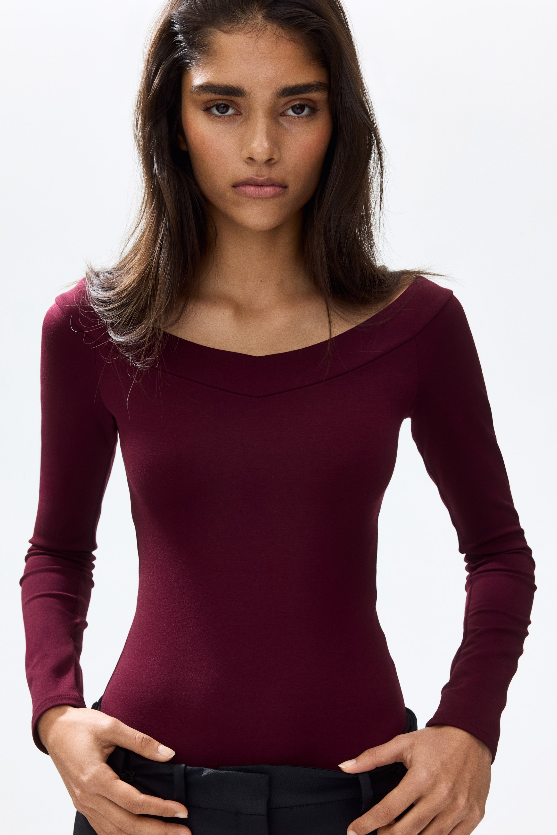 View larger image: The model is wearing a deep red, long-sleeved, off-the-shoulder bodysuit crafted from heavy Ponte di Roma jersey. It features a softly V-shaped, double-layered neckline. Her hands rest at her waist, where the bodysuit is neatly tucked into black trousers.