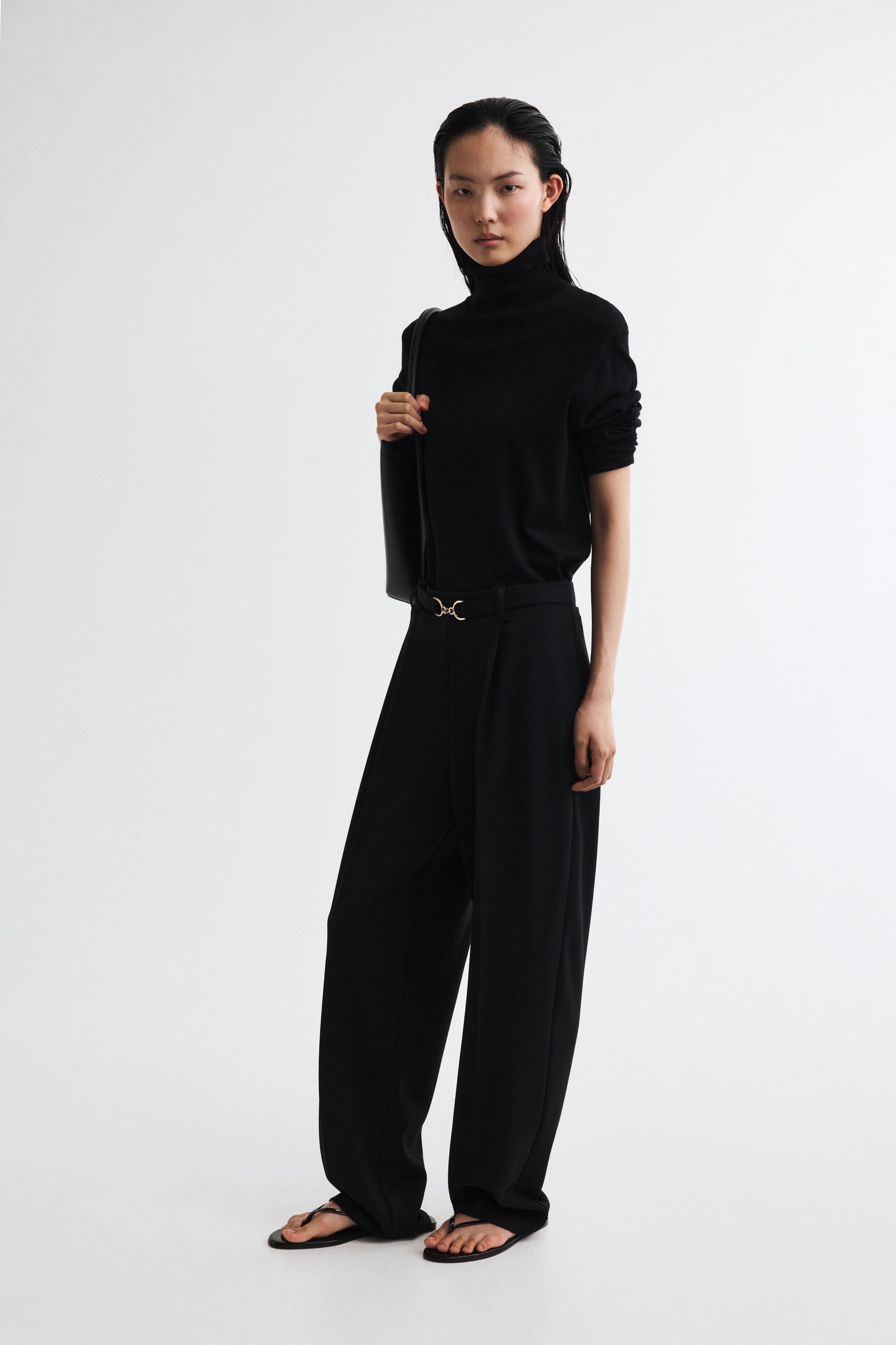 Büyük resmi görüntüle: Modelin üzerinde, ponte kumaştan üretilmiş siyah relaxed-fit pantolonlar yer alıyor; düz paçalı, üst pileli, ön ütü çizgili ve bel kısmında altın tonlu snaffle detaylı dekoratif bir kemer bulunuyor. Pantolonlar, içine sokulmuş siyah balıkçı yaka kazak ve siyah düz sandaletlerle kombinlenmiş.
