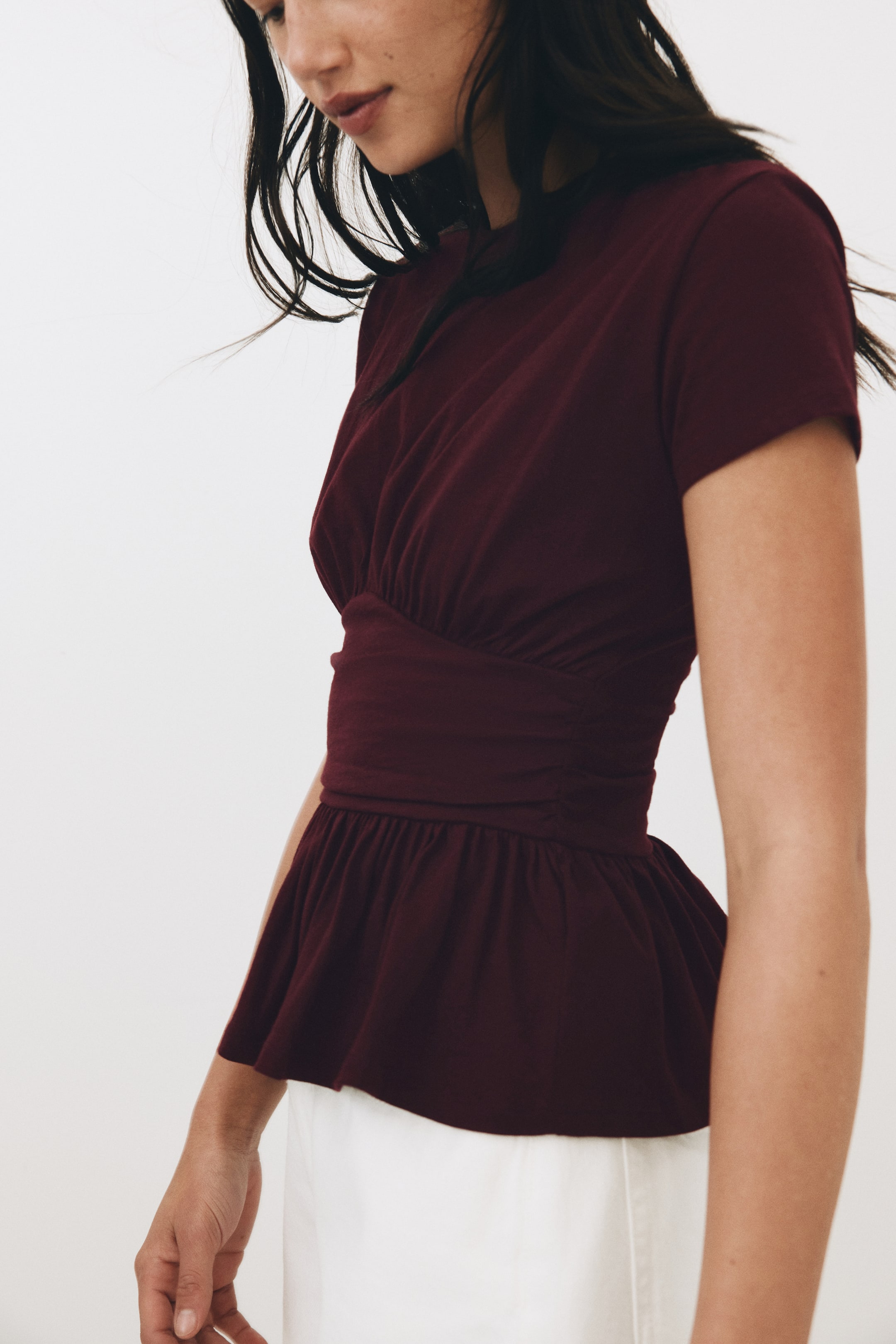 View larger image: The burgundy peplum top is shown from a slight side-front angle, highlighting its short sleeves and round neckline. It features a ruched bodice that transitions into a wide, cinched waistband, from which a gathered peplum hem flares out.