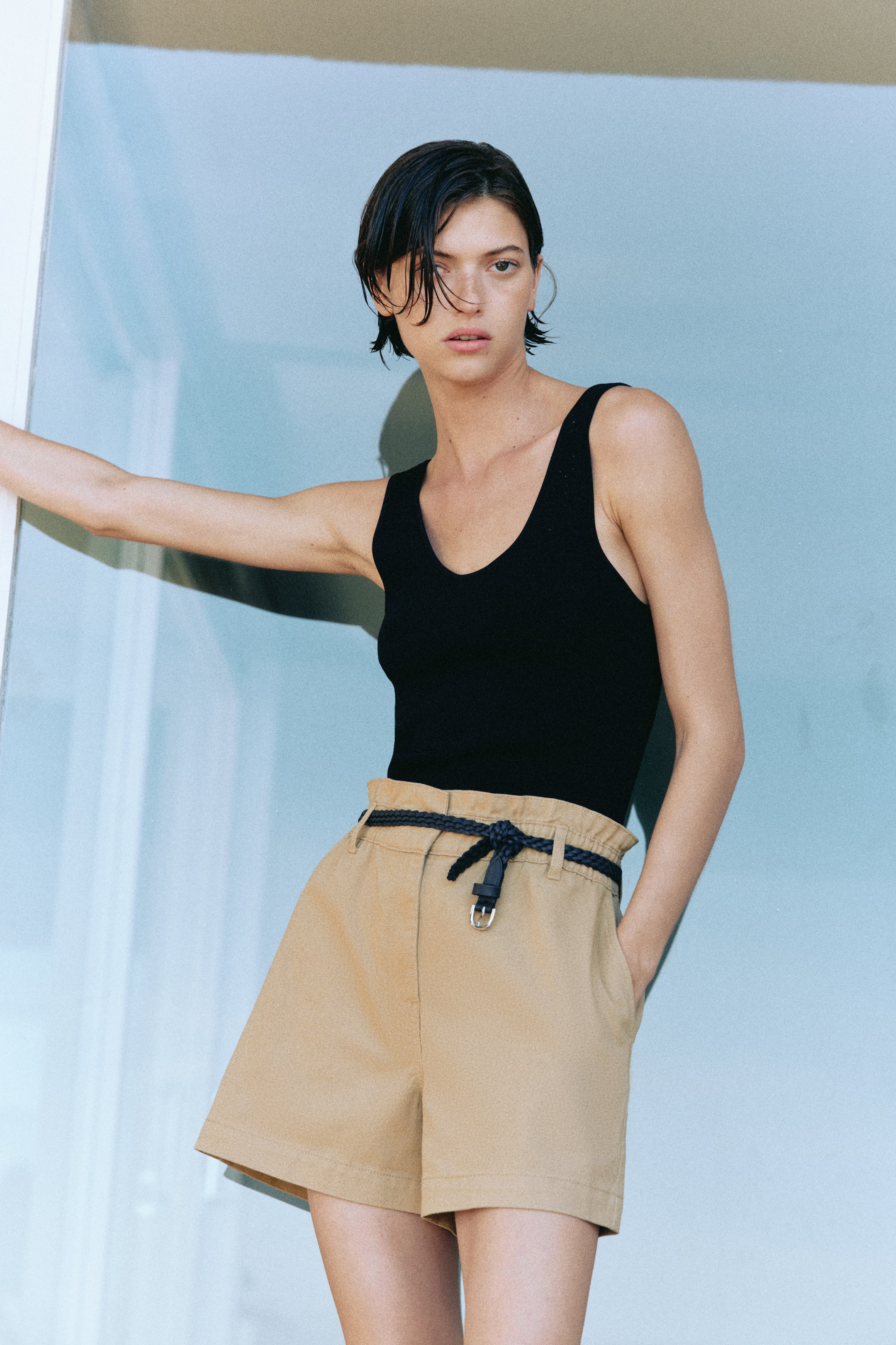 Grotere afbeelding bekijken: Een zwarte V-hals tanktop past strak. Lichtbruine shorts met hoge taille hebben een paperbag taille, een losse pasvorm en zijzakken, en zijn gestyled met een donkere gevlochten riem die in een knoop is vastgemaakt.