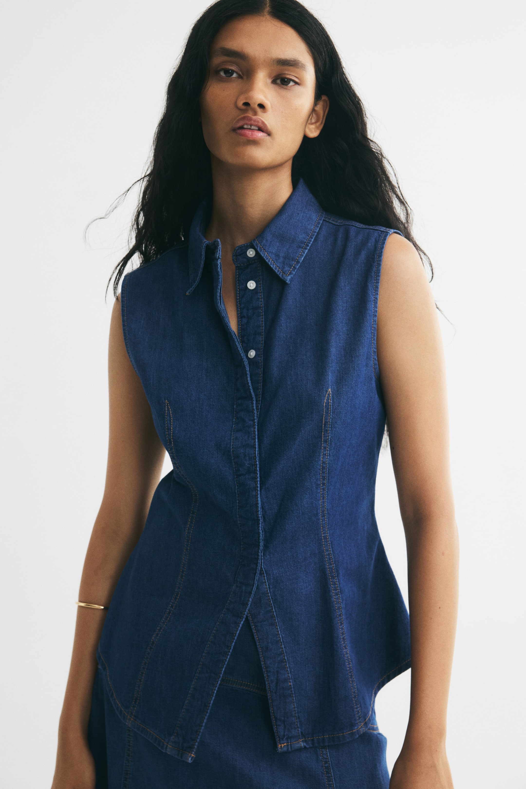 View larger image: The model is wearing a sleeveless dark blue denim shirt featuring a classic collar, a button-down front, and vertical shaping seams with contrast stitching. The slim-fit shirt has a slightly flared, rounded hem and is paired with a matching denim skirt.