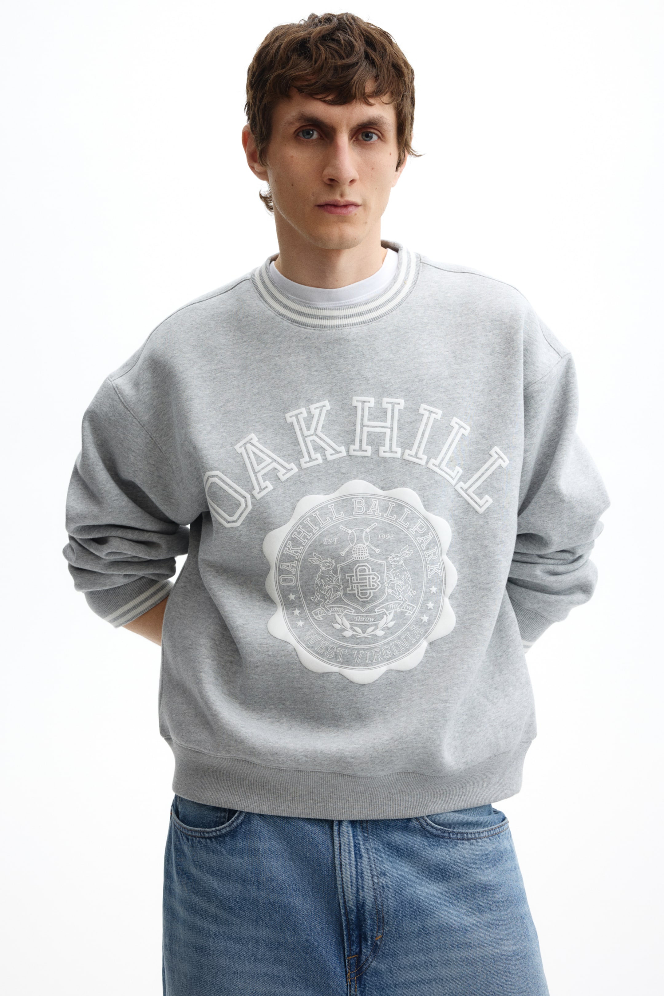 View larger image: A grey crewneck sweatshirt features a large white graphic print on the front. The print reads "Oakhill" in arched letters above a circular emblem detailing "Oakhill Ballpark West Virginia" and a crest motif. The sweatshirt is finished with ribbed cuffs and hem and a striped ribbed collar, and is shown layered over a white T-shirt.