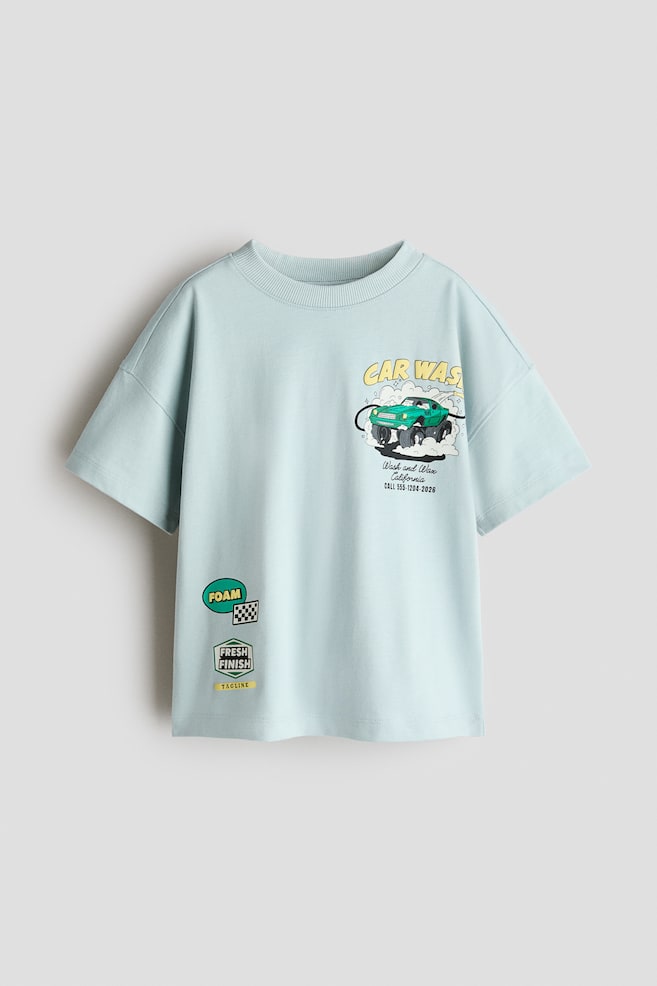 A light blue, oversized t-shirt is facing front, featuring a ribbed crewneck, dropped shoulders, and short sleeves. The front displays a colorful car wash graphic on the chest and smaller, patch-like graphic prints on the lower side. - 1