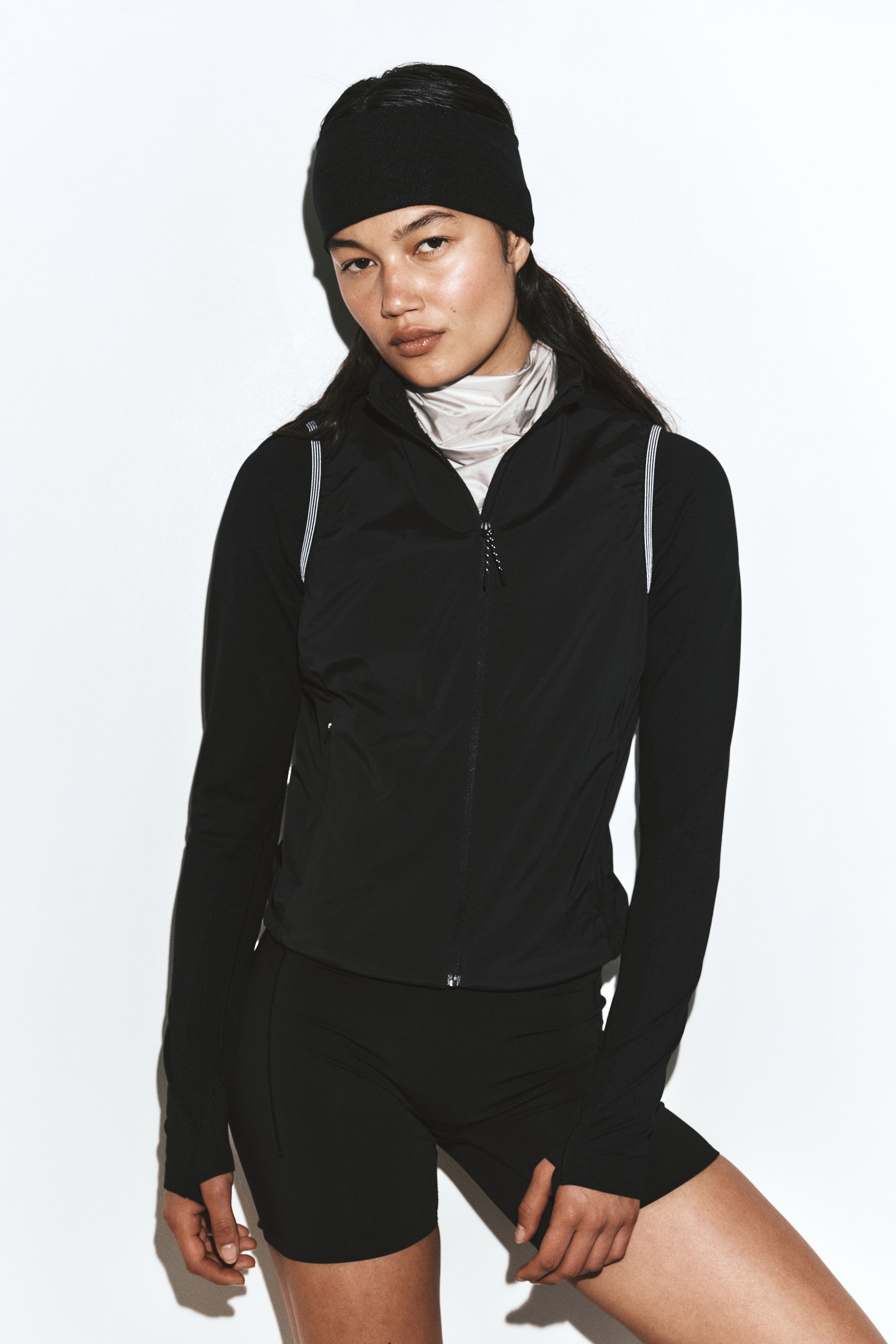 View larger image: A black, slim-fit activewear vest is being worn by the model over a black long-sleeve top and matching athletic shorts. The vest features a stand collar, a centre front zipper, and thin reflective piping along the armholes.