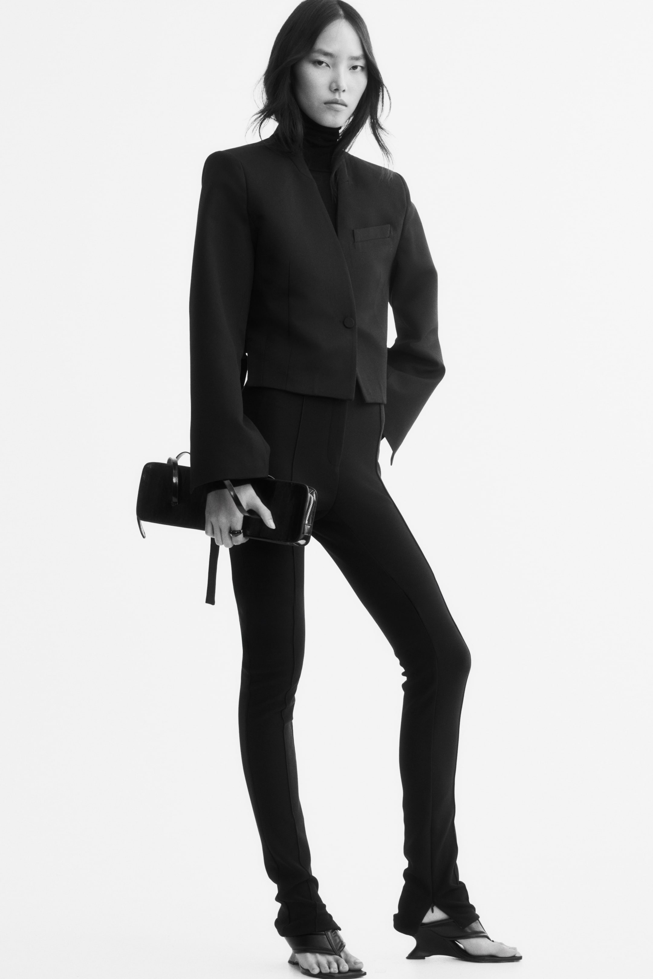 Agrandir l'image: Un legging noir en jersey extensible, doté de plis cousus sur le devant, est porté par le mannequin. Ce legging slim fit est associé à un blazer noir court, à un haut à col montant et à des sandales à talons.