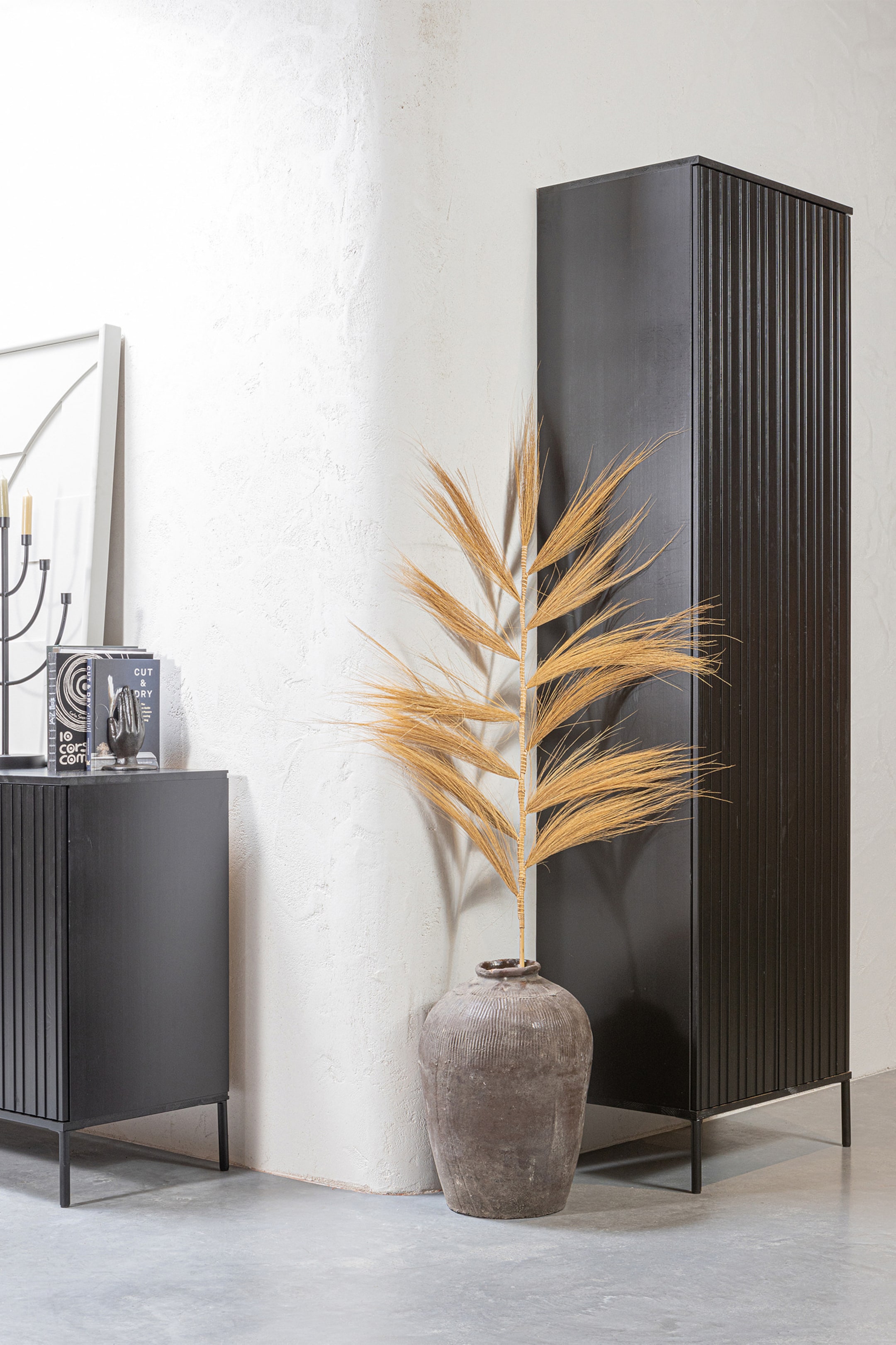 Größeres Bild ansehen: Ein hoher schwarzer Holzschrank und ein passendes niedriges Sideboard stehen auf schlanken Metallbeinen und zeigen vertikale gerillte Linienmuster über ihre grifflosen Fronttüren. Der hohe Schrank ist in einer Dreiviertelansicht positioniert, um sowohl seine glatte Seitenwand als auch die strukturierte Vorderseite hervorzuheben.