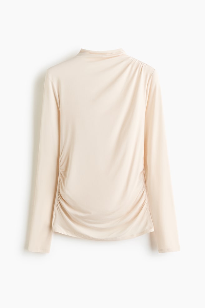 A light beige long-sleeved top with a mock neck and gentle ruching on the left shoulder and along the left side seam, creating elegant drapes across the back. - 1