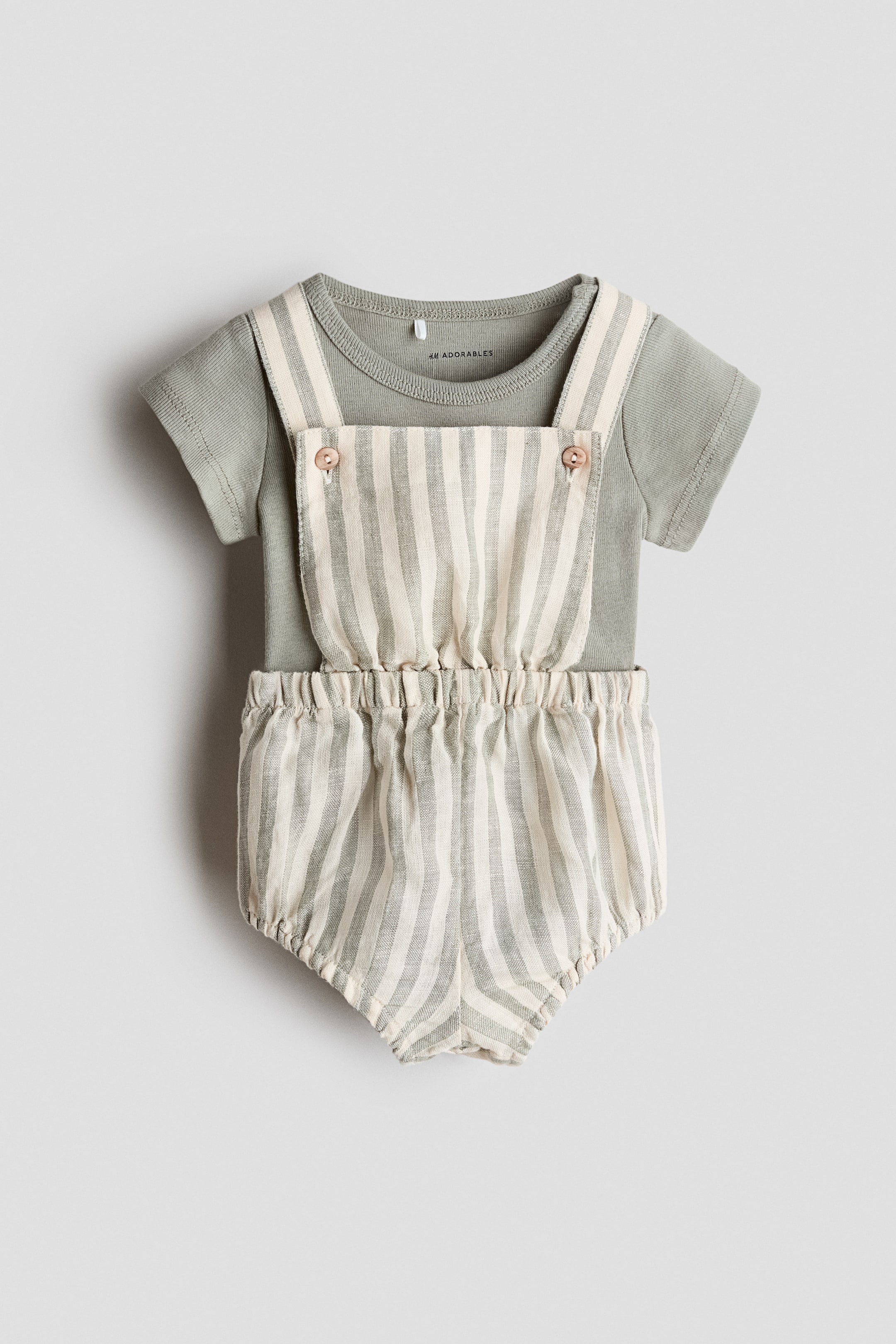 Agrandir l'image: Une tenue pour bébé est présentée de face, comprenant un haut à manches courtes vert clair avec un col rond côtelé. Par-dessus, un barboteuse affiche des rayures verticales vert clair et crème, avec des bretelles se fermant par deux boutons marron clair sur la bavette avant, ainsi qu'une taille et des ouvertures de jambes élastiquées et froncées.