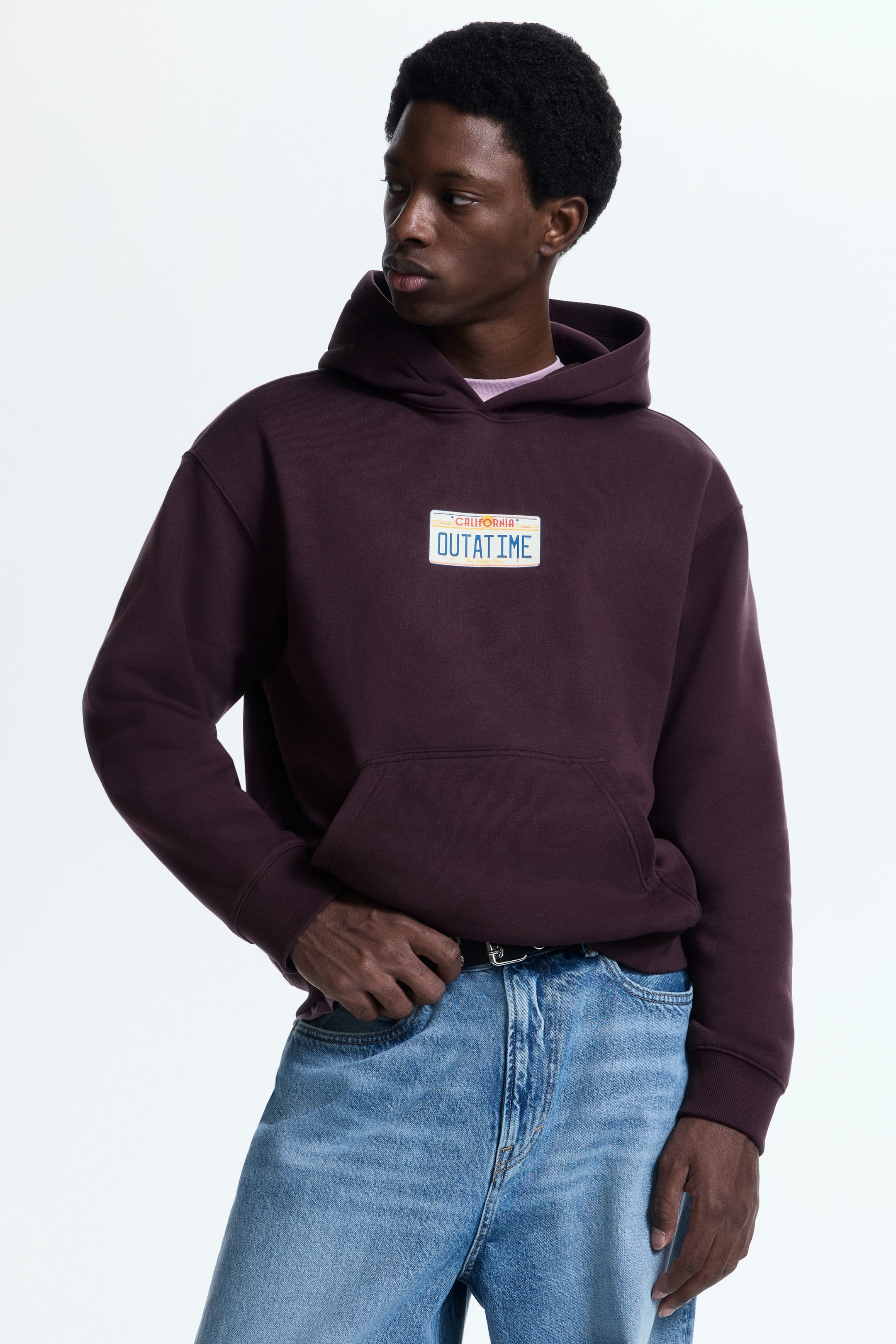 View larger image: A maroon oversized hoodie is facing slightly to the right, displaying a rectangular license plate graphic on the chest with the text 'California' and 'OUTATIME'. It is featuring an attached hood, a front kangaroo pocket, and ribbed cuffs and hem, with a light pink t-shirt visible underneath.