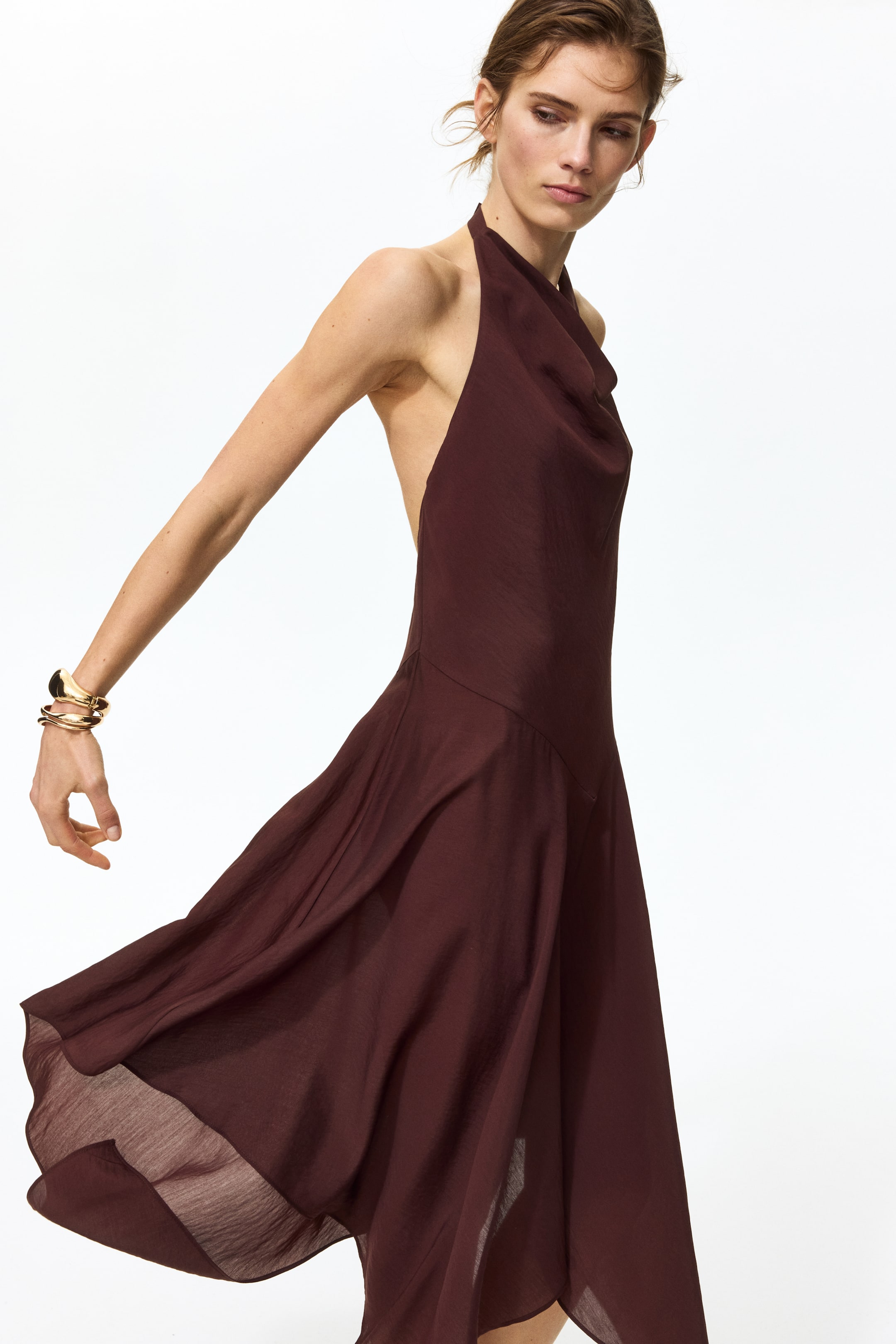 View larger image: A burgundy halter neck dress featuring a draped cowl neckline and a low-cut back. The lightweight fabric of the flowing skirt moves outward, creating soft folds.
