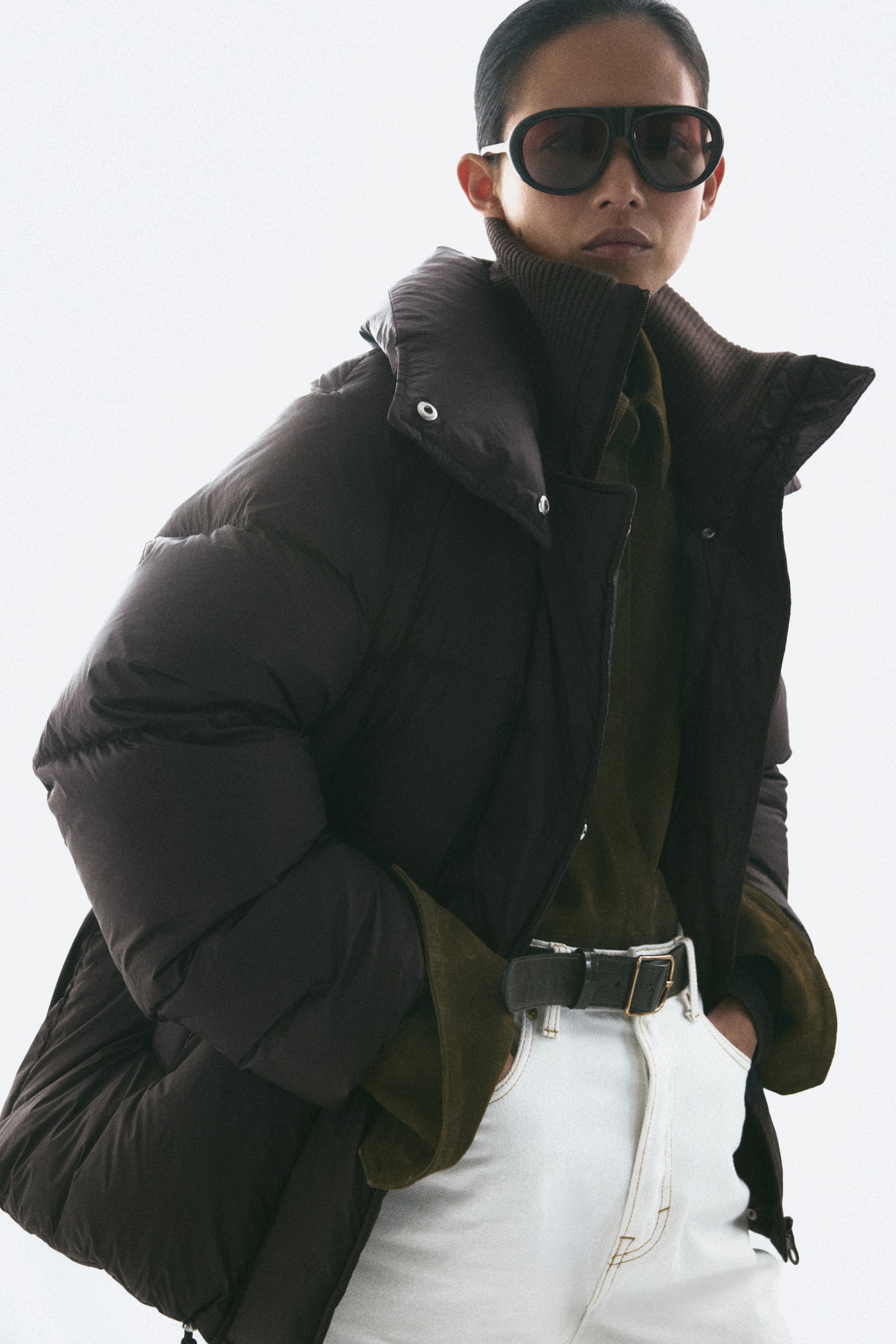 A dark brown puffer jacket is shown from the front, featuring a quilted design with a double-layered funnel collar and a ribbed inner collar. The jacket is displaying raglan sleeves with ribbed inner cuffs, diagonal welt pockets, and drawstrings hanging from the hem. - 1