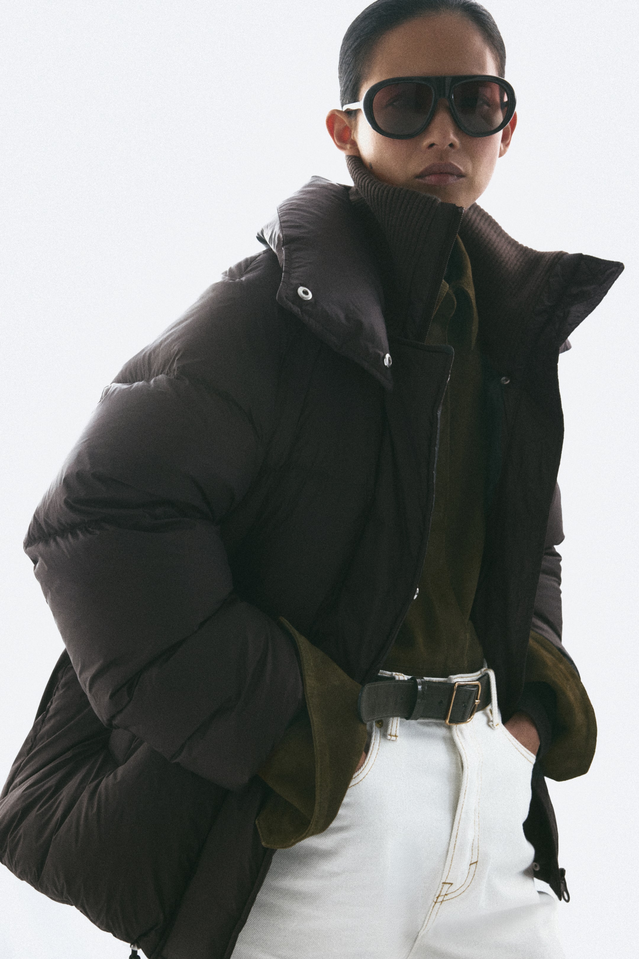 View larger image: A dark brown padded nylon jacket is being worn by the model, featuring a high, double-layered funnel collar with a ribbed inner collar and voluminous quilted sections. The jacket is styled open, layered over a dark green collared shirt and white jeans.