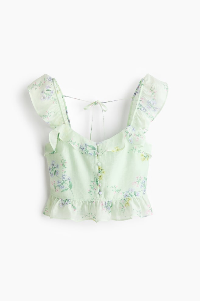 A light green floral crop top is showcased front-facing, featuring a ruffled sweetheart neckline and wide ruffled shoulder straps. It is designed with a button-through front fastening, a ruffled peplum hem, and a tie-back detail at the upper back. - 1