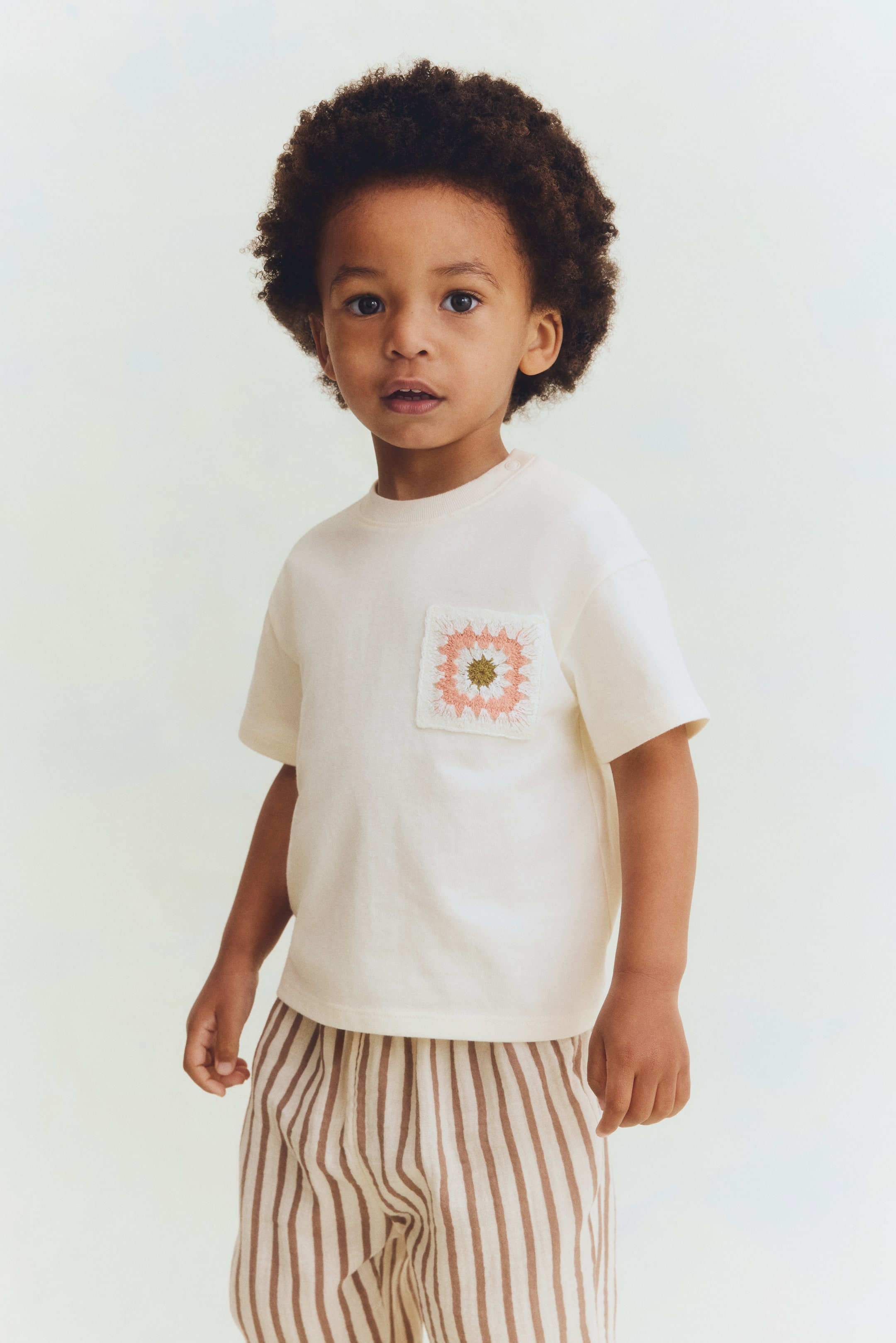Visualizza l'immagine ingrandita: Una t-shirt color crema presenta un collo rotondo, maniche corte e due bottoni a pressione sulla spalla sinistra, con una tasca quadrata all'uncinetto che mostra un design floreale bianco, rosa e verde sul petto sinistro. È abbinata a pantaloni di colore chiaro con strisce verticali marroni.