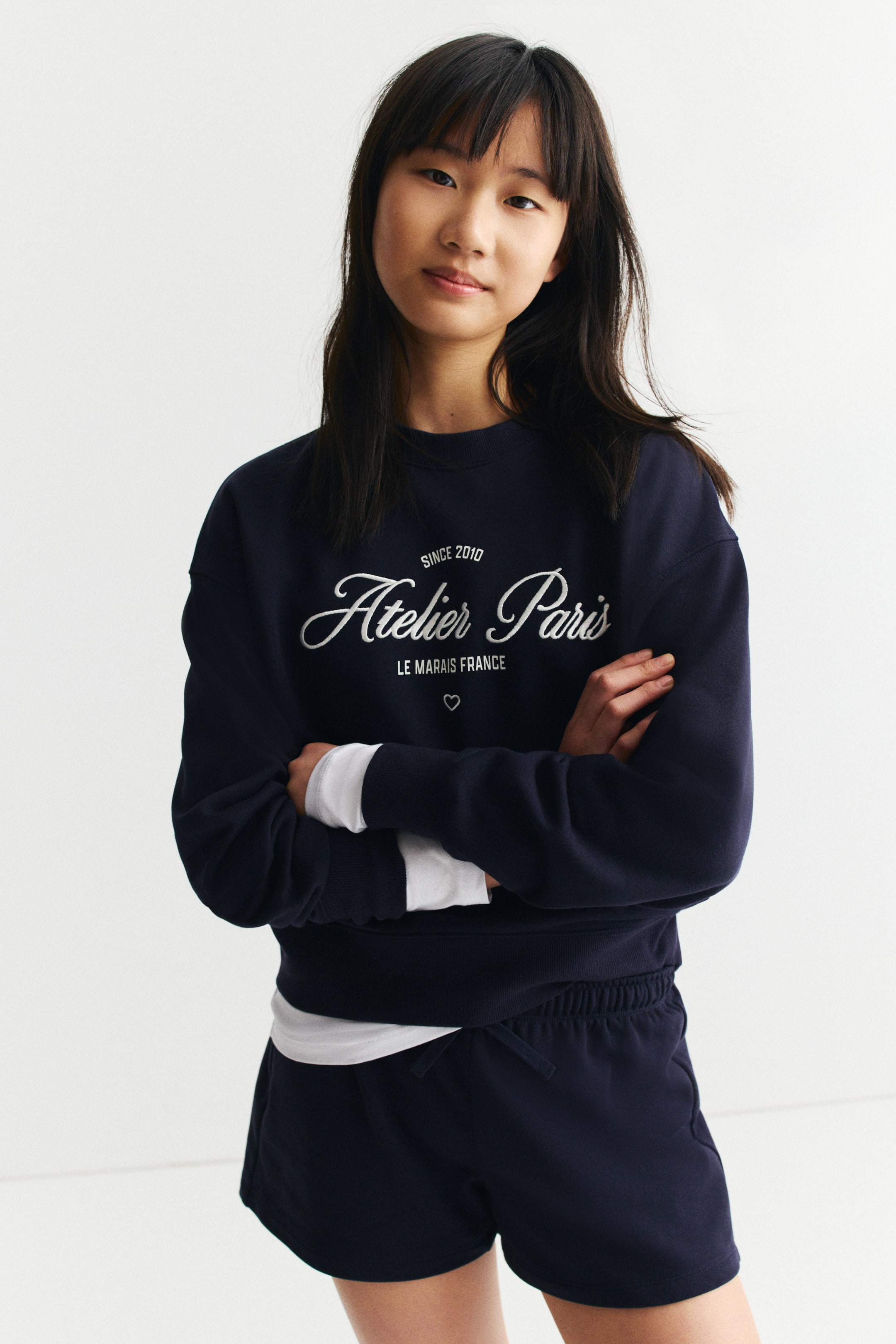 Grösseres Bild ansehen: Ein marineblaues Sweatshirt mit Rundhalsausschnitt zeigt den weißen Text "SINCE 2010 Atelier Paris LE MARAIS FRANCE" mit einem Herzsymbol auf der Vorderseite, gerippte Bündchen und einen gerippten Saum. Es wird über einem weißen langärmligen Hemd getragen und mit passenden marineblauen Shorts kombiniert, die über einen elastischen Kordelzugbund und Seitentaschen verfügen, alles in frontaler Ansicht präsentiert.