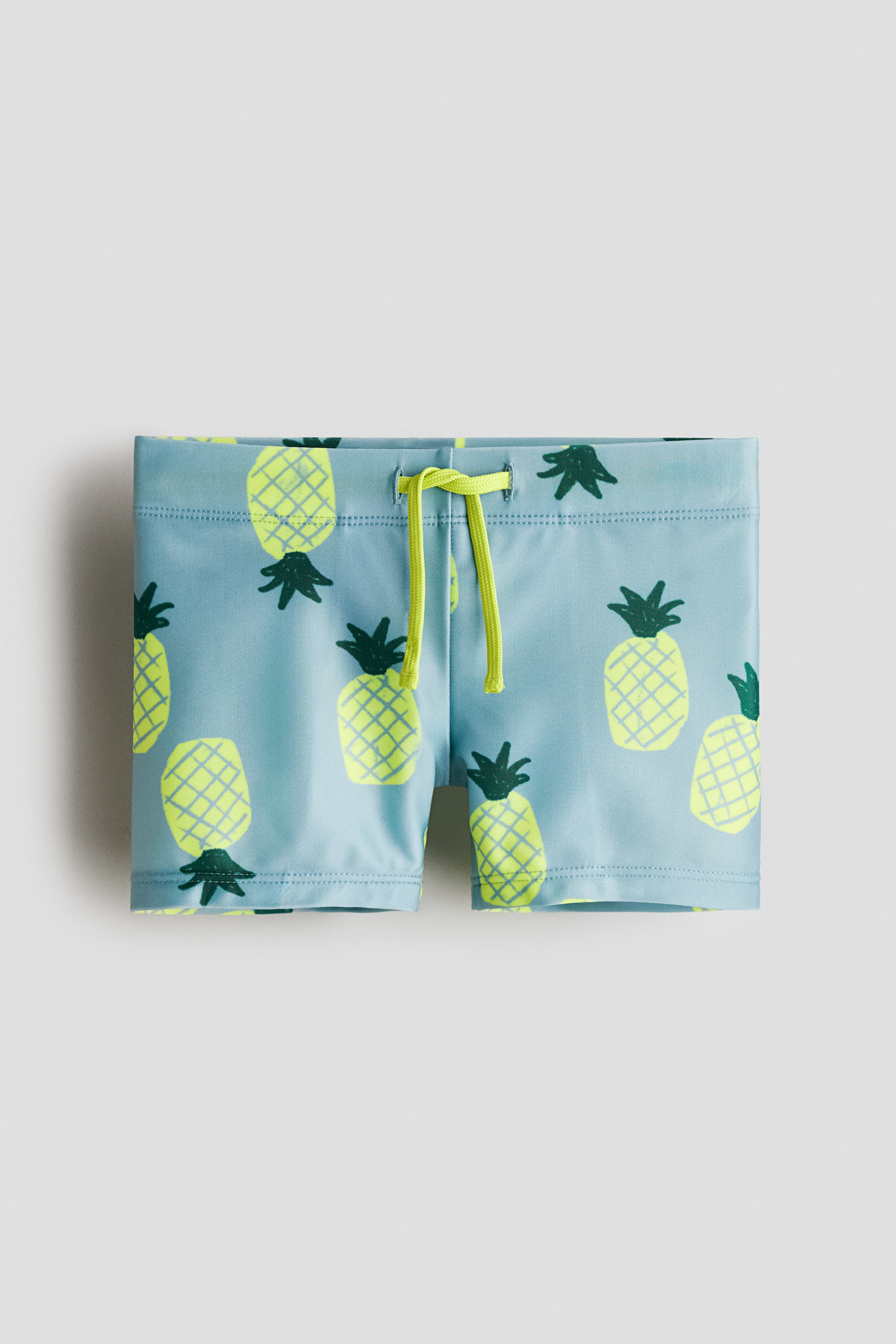 Prikaži veću sliku: Šorts za plivanje prikazuju prednji pogled, sa svetloplavim platnom ukrašenim ponavljajućim žutim uzorkom ananasa. Dizajnirani su sa elastičnim pojasom na vezivanje, gde je svetložuti konopac zavezan na sredini prednjeg dela.