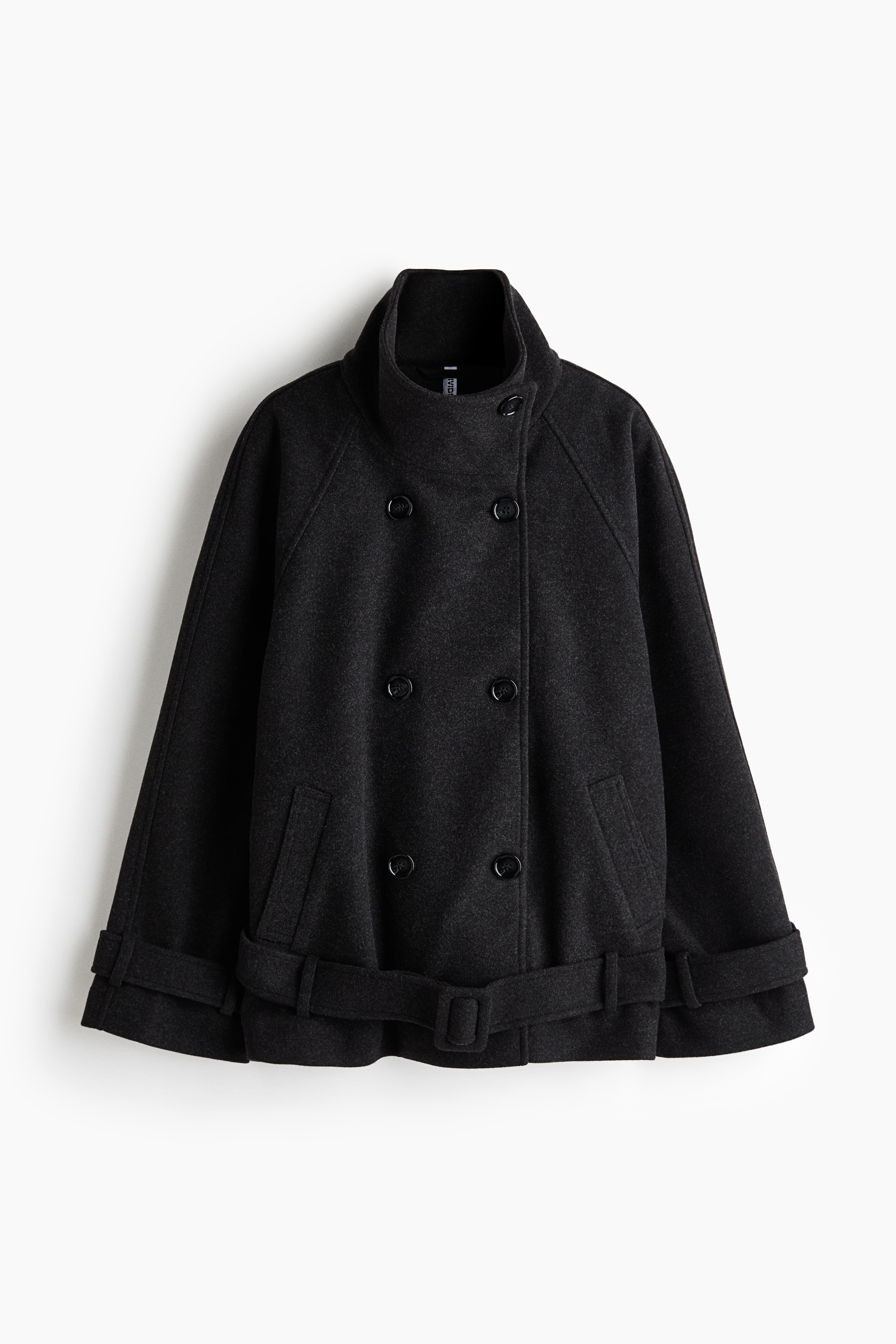 An oversized, double-breasted jacket in dark grey felted fabric is being worn by the model, who is resting their hands inside the front welt pockets. The jacket features a high buttoned funnel collar, raglan sleeves with button-tab cuffs, and an adjustable matching belt at the hem.