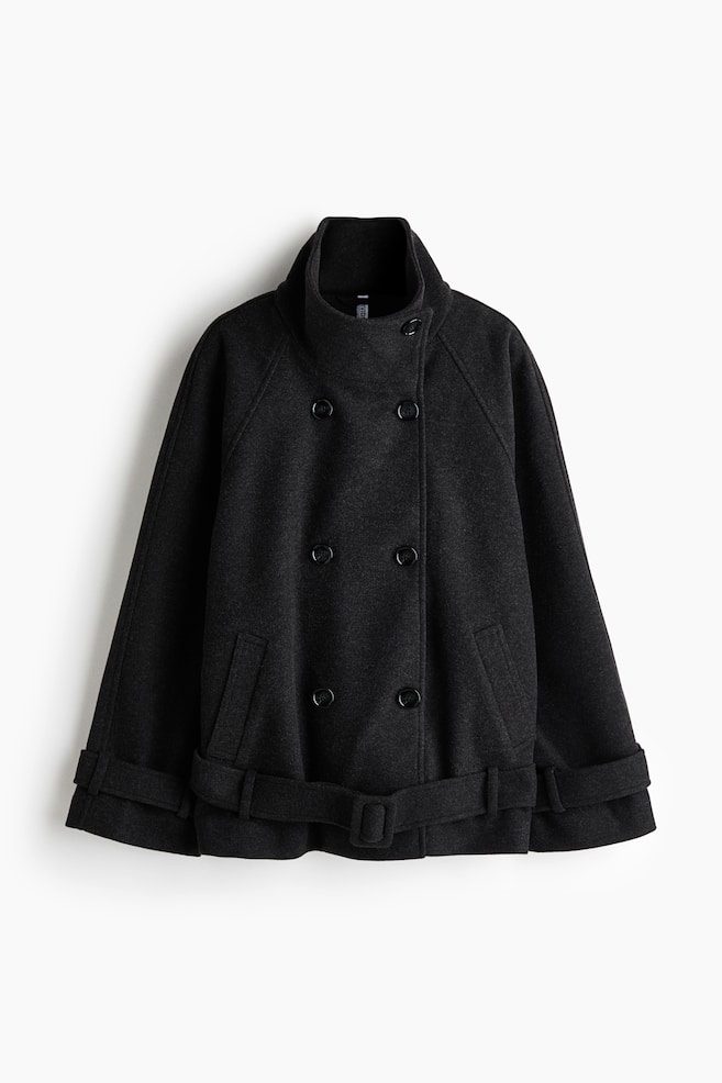 An oversized, double-breasted jacket in dark grey felted fabric is shown from the front, featuring a buttoned funnel collar, raglan sleeves with button-tab cuffs, and angled welt front pockets. The jacket is styled with a matching adjustable belt containing a covered buckle at the hem. - 1
