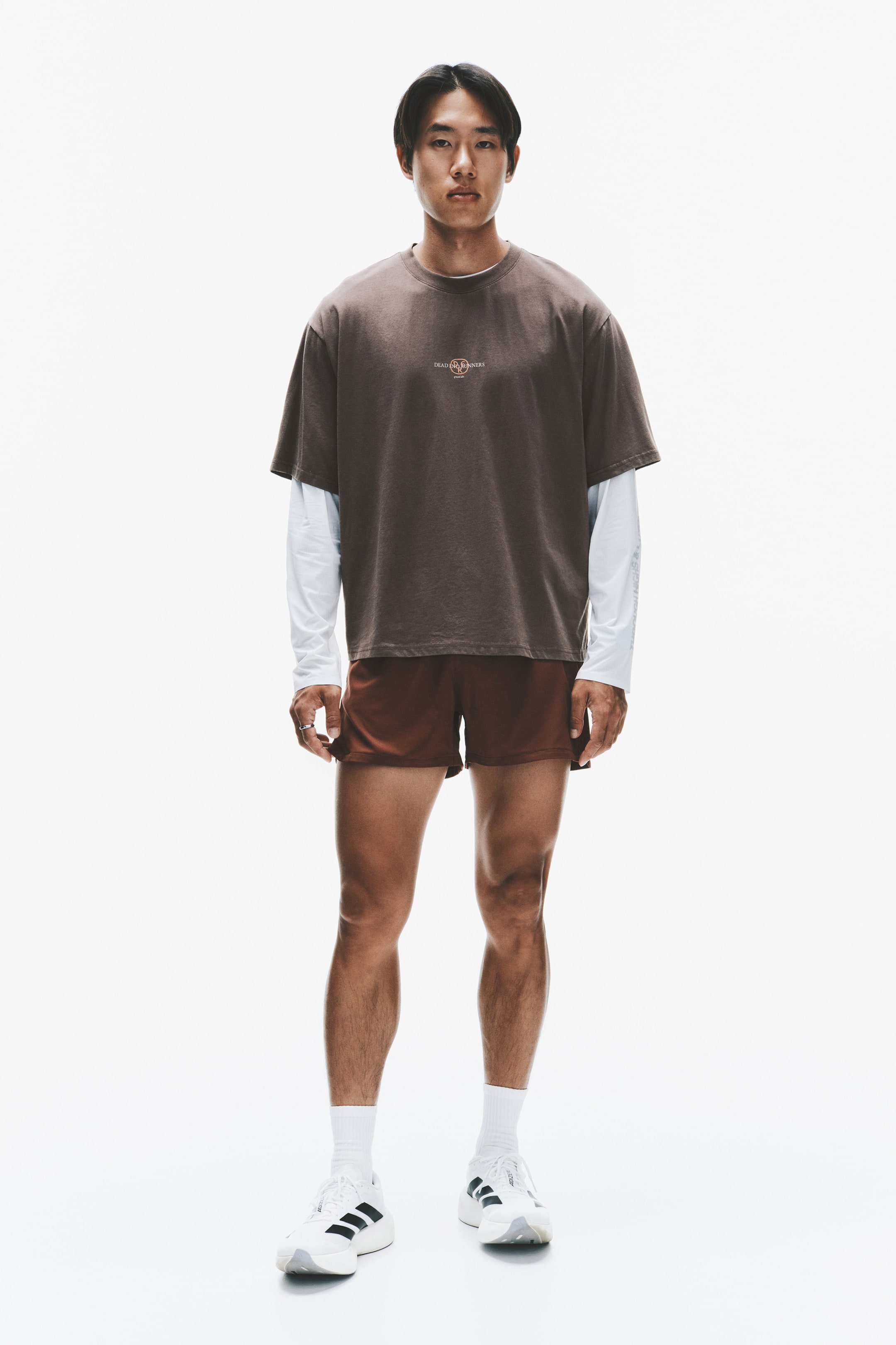 View larger image: A dark brown, oversized short-sleeved T-shirt featuring a small central chest graphic is layered over a white long-sleeved shirt, paired with brown athletic shorts. The outfit is shown from a front-facing angle, highlighting the relaxed fit of the top and the mid-thigh length of the shorts.