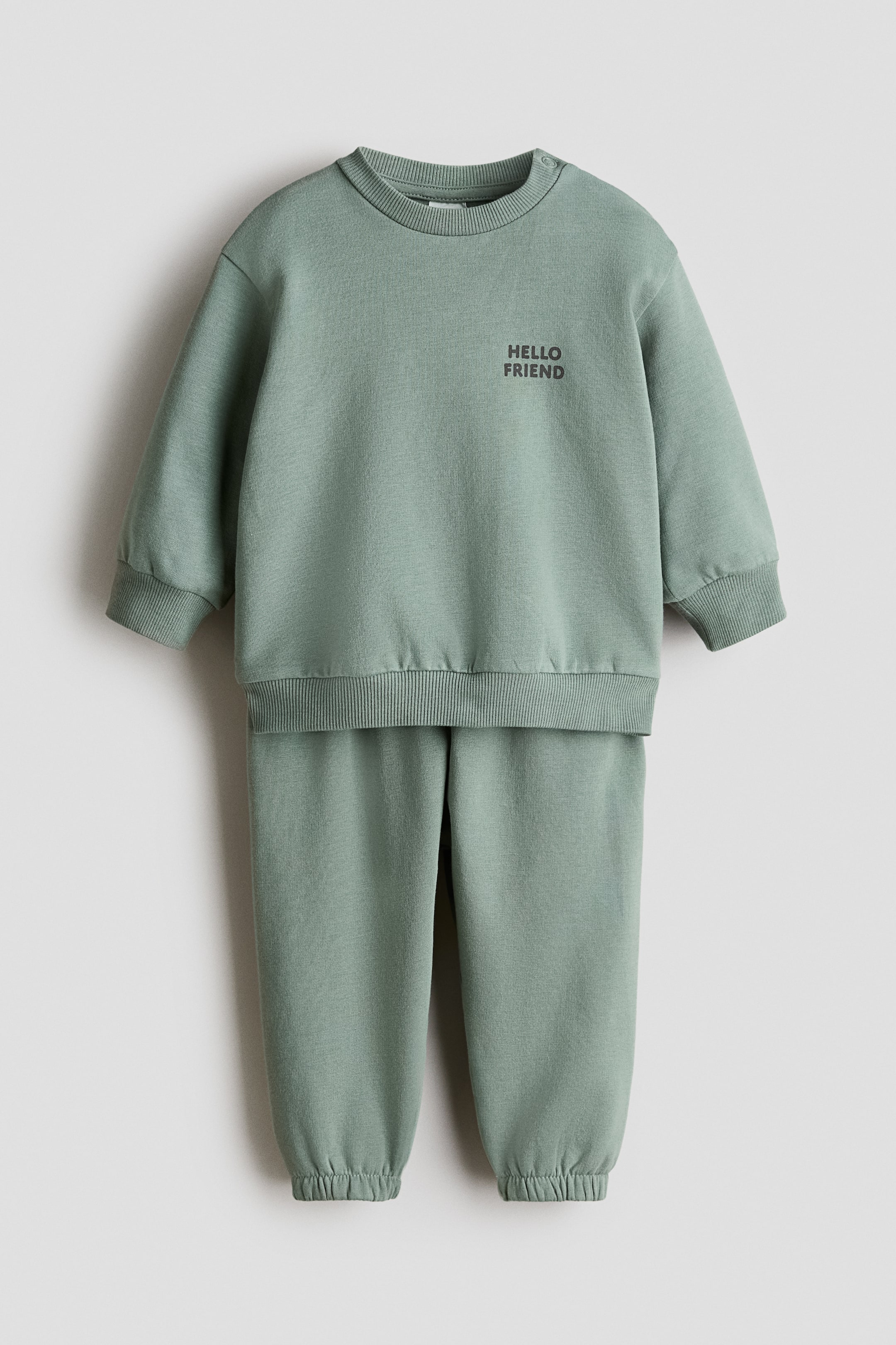 View larger image: A light green sweatshirt and tracksuit bottoms set is displaying a front view, laid flat. The sweatshirt is featuring a ribbed crew neck with two snap buttons on the left shoulder, ribbed cuffs, a ribbed hem, and "HELLO FRIEND" printed on the chest. The matching tracksuit bottoms are completing the set with elasticated cuffs.
