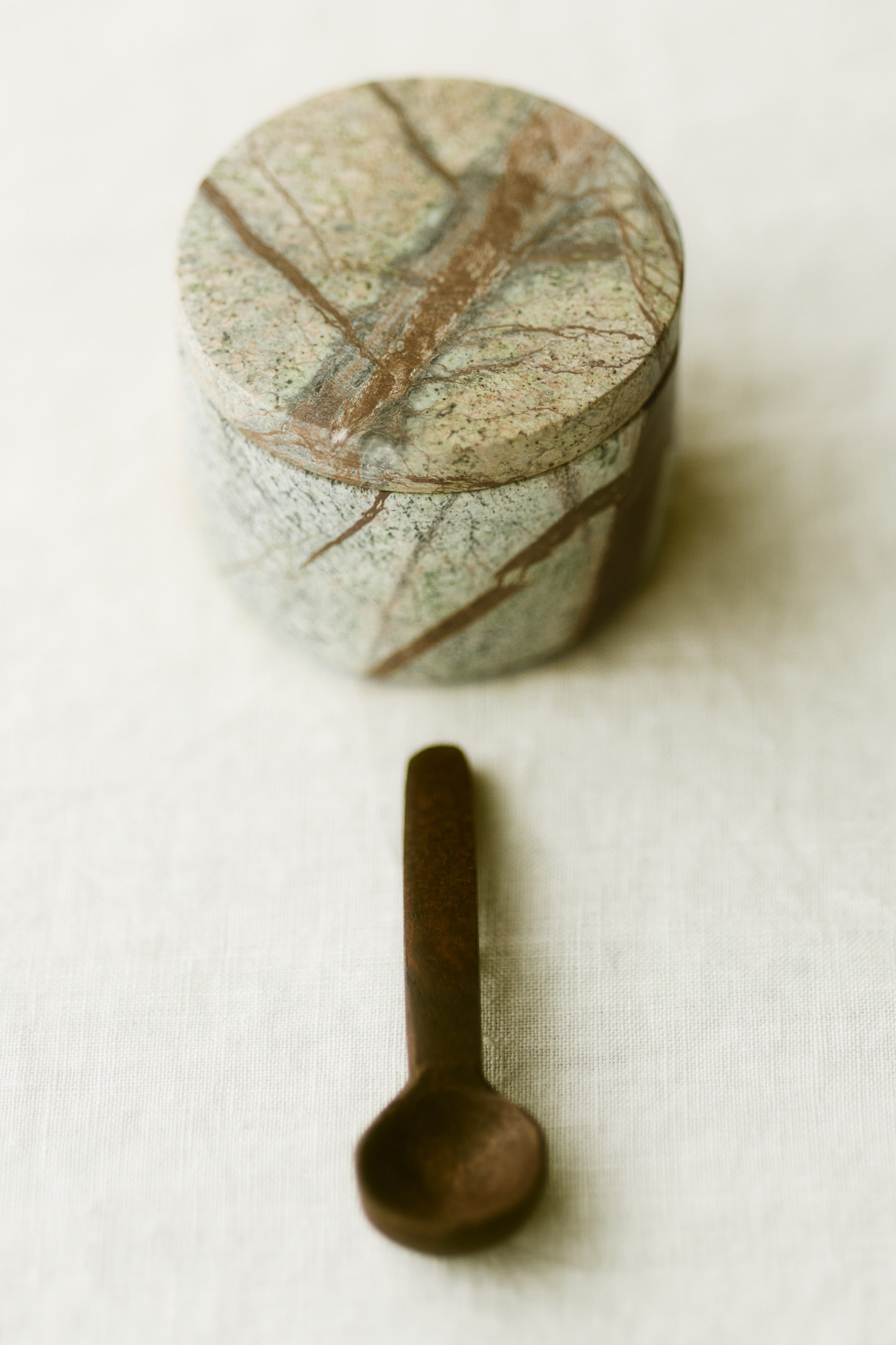 View larger image: A cylindrical container made from green and brown veined marble features a flat lid on its top surface, with a small, dark brown wooden spoon lying flat beside it.