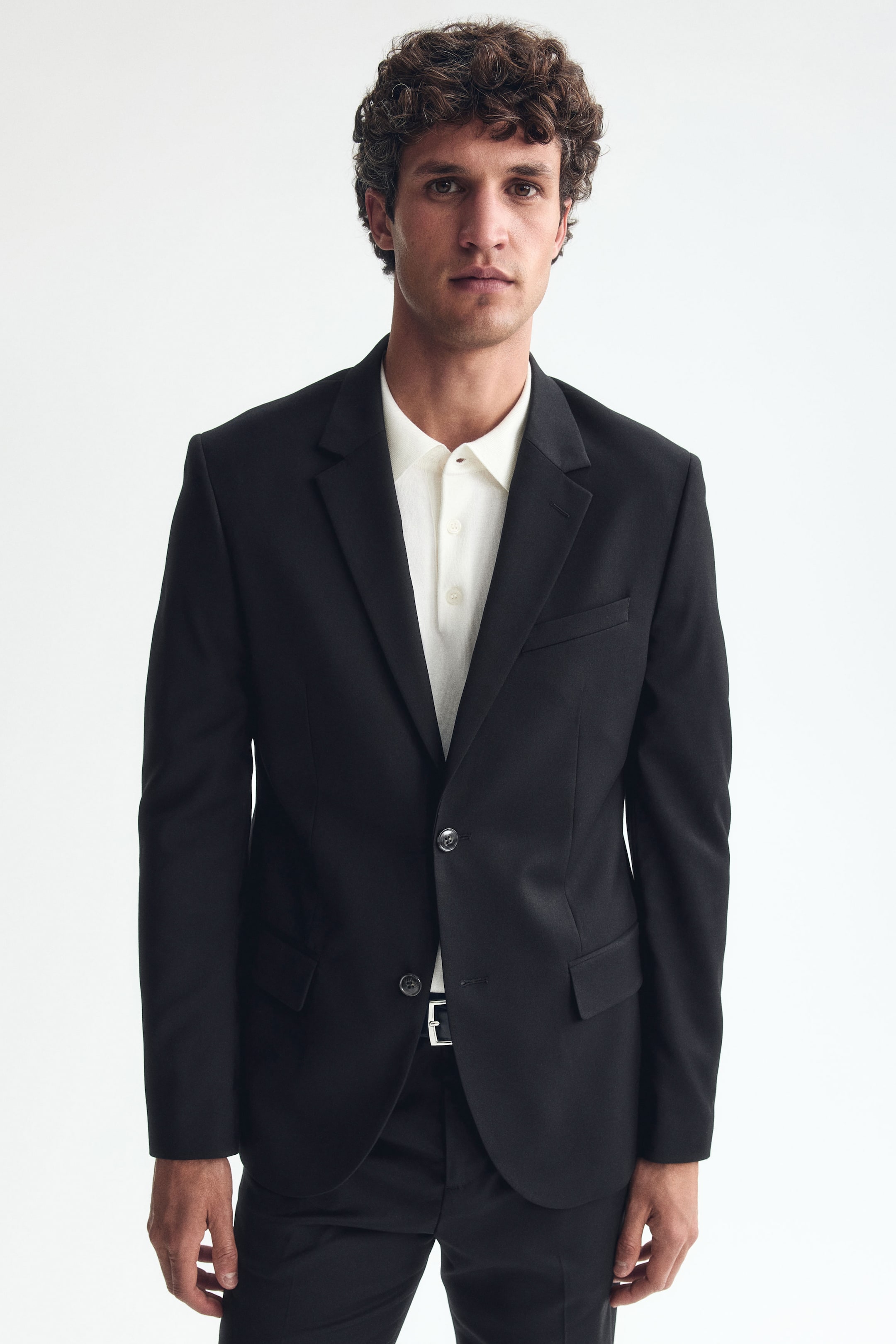 View larger image: The model is wearing a black single-breasted jacket, buttoned at the front. It showcases notch lapels, a jetted chest pocket, and two flap front pockets, forming a fitted silhouette over a light-coloured polo shirt.