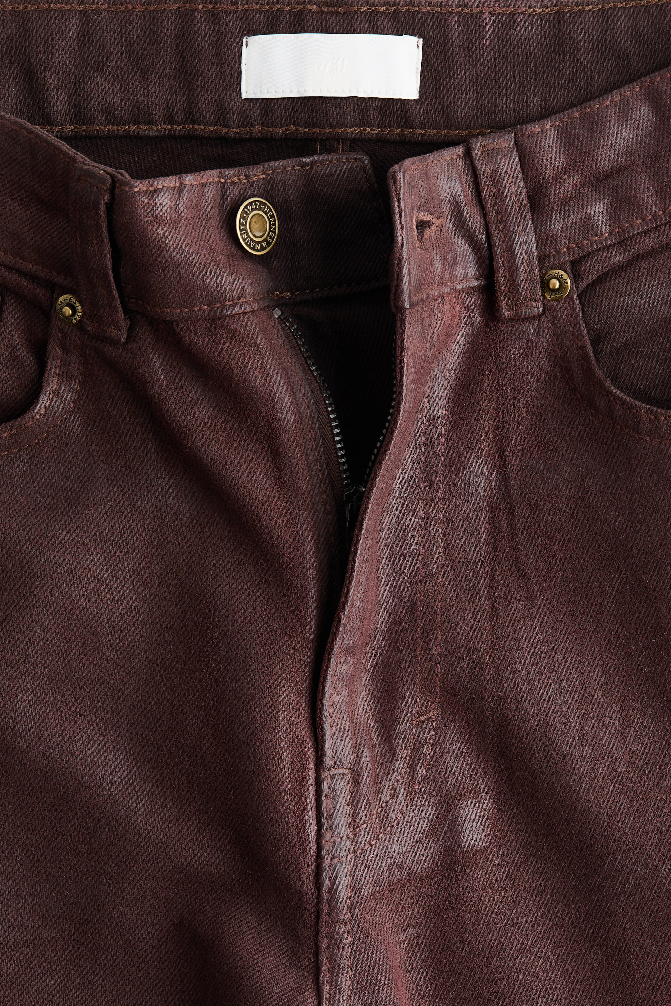 Visualizza immagine più grande: I jeans marrone scuro frontali presentano una leggera lucentezza, una cerniera parzialmente aperta e una chiusura con un bottone di metallo alla vita. I jeans mostrano anche passanti per cintura, rivetti di metallo sulle tasche anteriori e un'etichetta bianca all'interno della cintura.