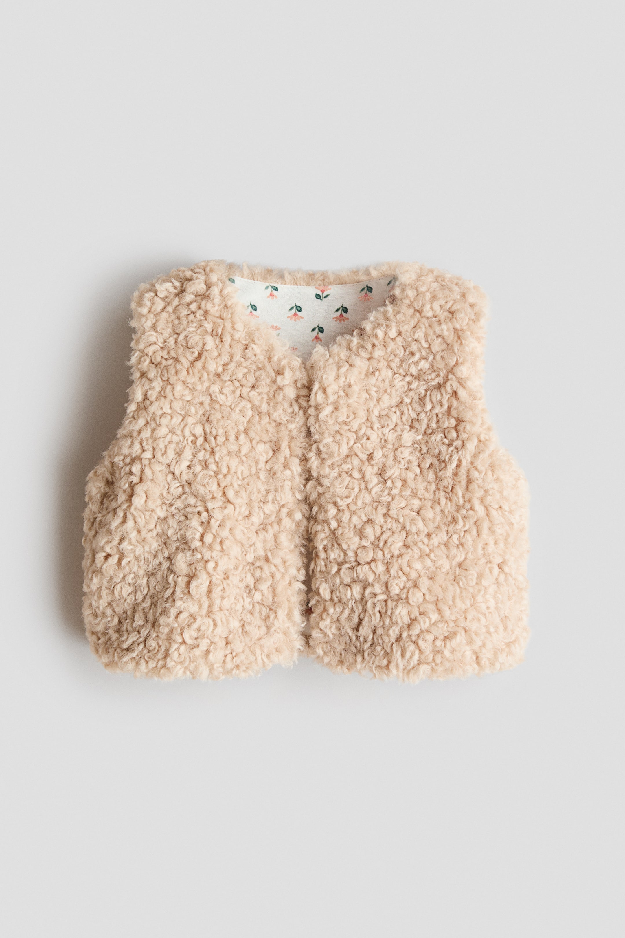 View larger image: A light beige faux fur vest showcases a curly texture, featuring a V-neckline and an open front. The vest reveals a white lining around the neckline, adorned with small pink and green floral patterns.