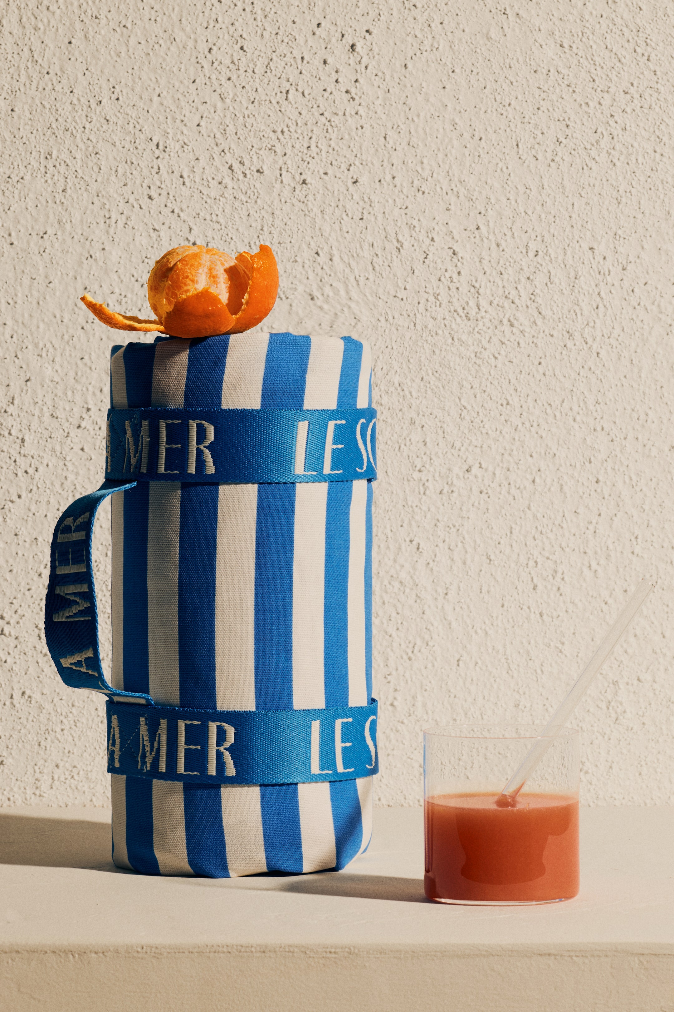 Ampliar la imagen: Un contenedor cilíndrico muestra rayas verticales azules y blancas, y correas de tela azul con texto blanco envuelven su cuerpo. Una de las correas forma un asa en su lado izquierdo, y una cáscara de naranja descansa en la parte superior del contenedor.