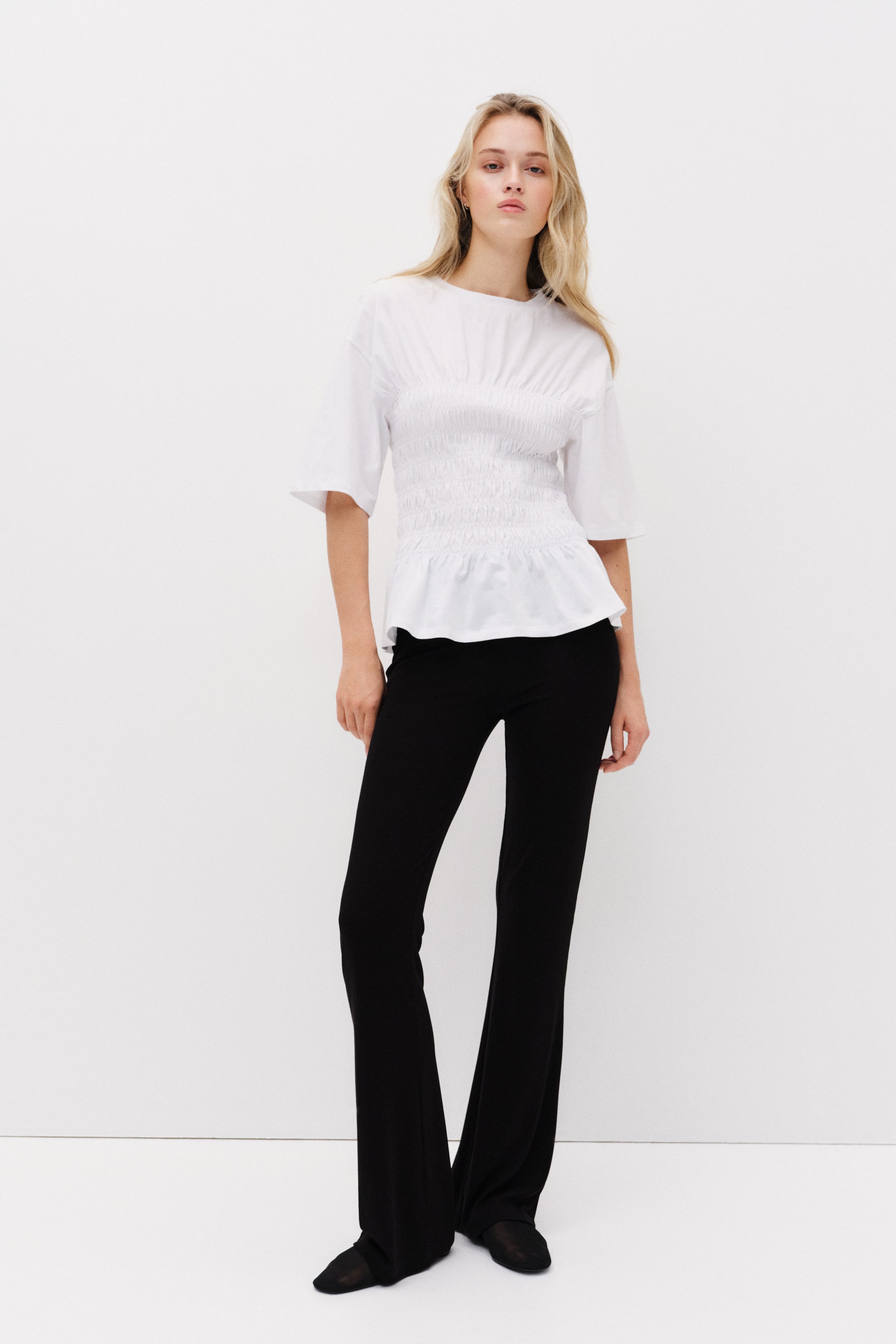 Zobacz większy obraz: Produkt to biały top i czarne spodnie, przedstawione przodem. Biały top charakteryzuje się okrągłym dekoltem, krótkimi szerokimi rękawami, mocno marszczonym korpusem i rozszerzającą się dolną częścią, podczas gdy czarne spodnie mają dopasowany krój w udach i rozszerzają się poniżej kolana do pełnej długości.