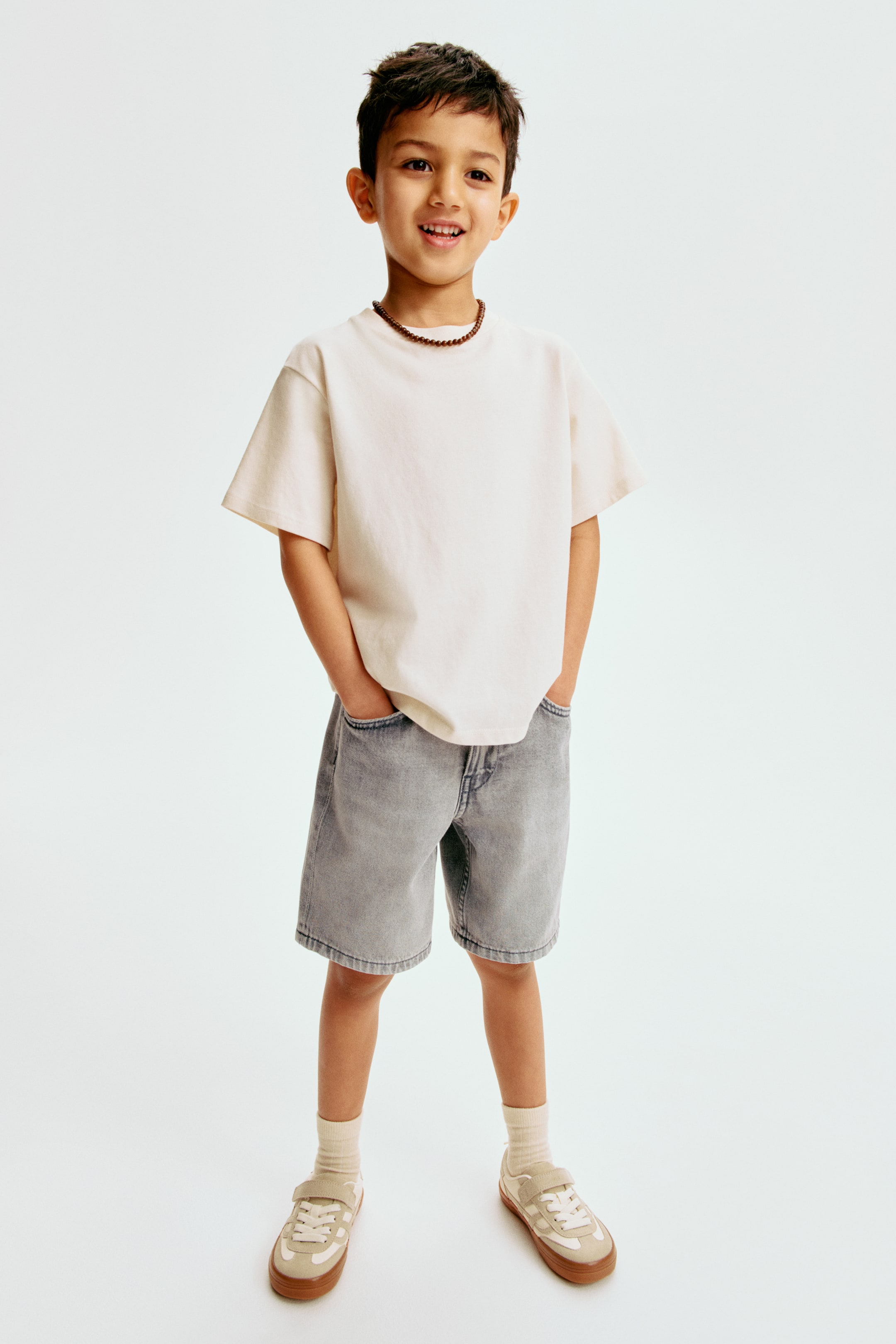 הצגת תמונה גדולה יותר: חולצת טי בצבע בז' בהיר עם צווארון עגול, שרוולים קצרים ורחבים ושוליים ישרים נלבשת עם מכנסי ג'ינס אפורים דהויים עם כיסים קדמיים גלויים. שרשרת חרוזים חומה מעטרת את הצוואר, בעוד גרביים בצבע בז' בהיר משתלבות עם נעלי סניקרס בצבע בז' ולבן-שבור עם סוליות חומות.