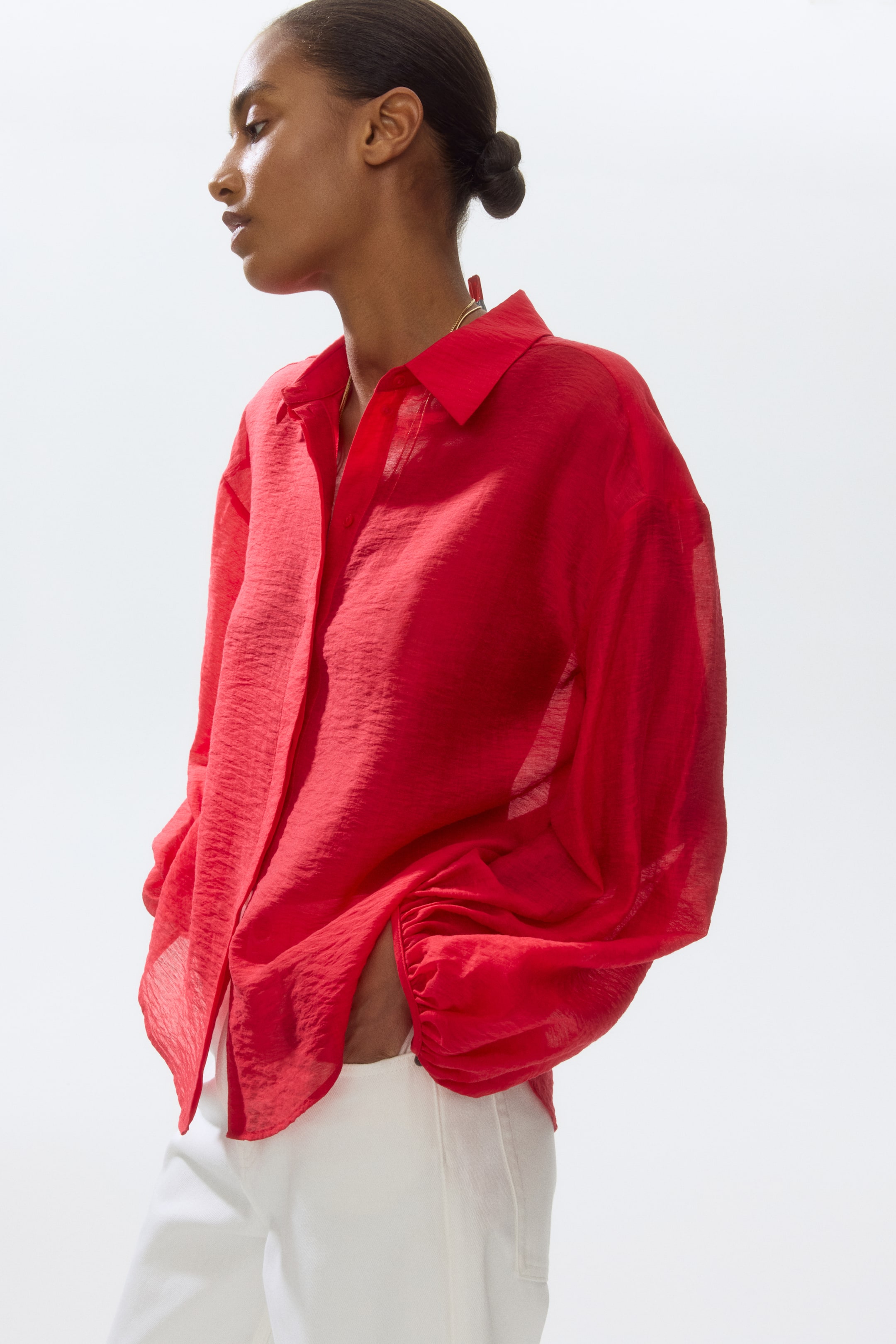 View larger image: A red textured button-up shirt is displayed from a side-front angle, highlighting its pointed collar, button-front closure, and long, voluminous sleeves gathered at elasticated cuffs. The shirt fits loosely, with its front hem pulled by the wearer's hands in pockets, emphasizing its relaxed drape.