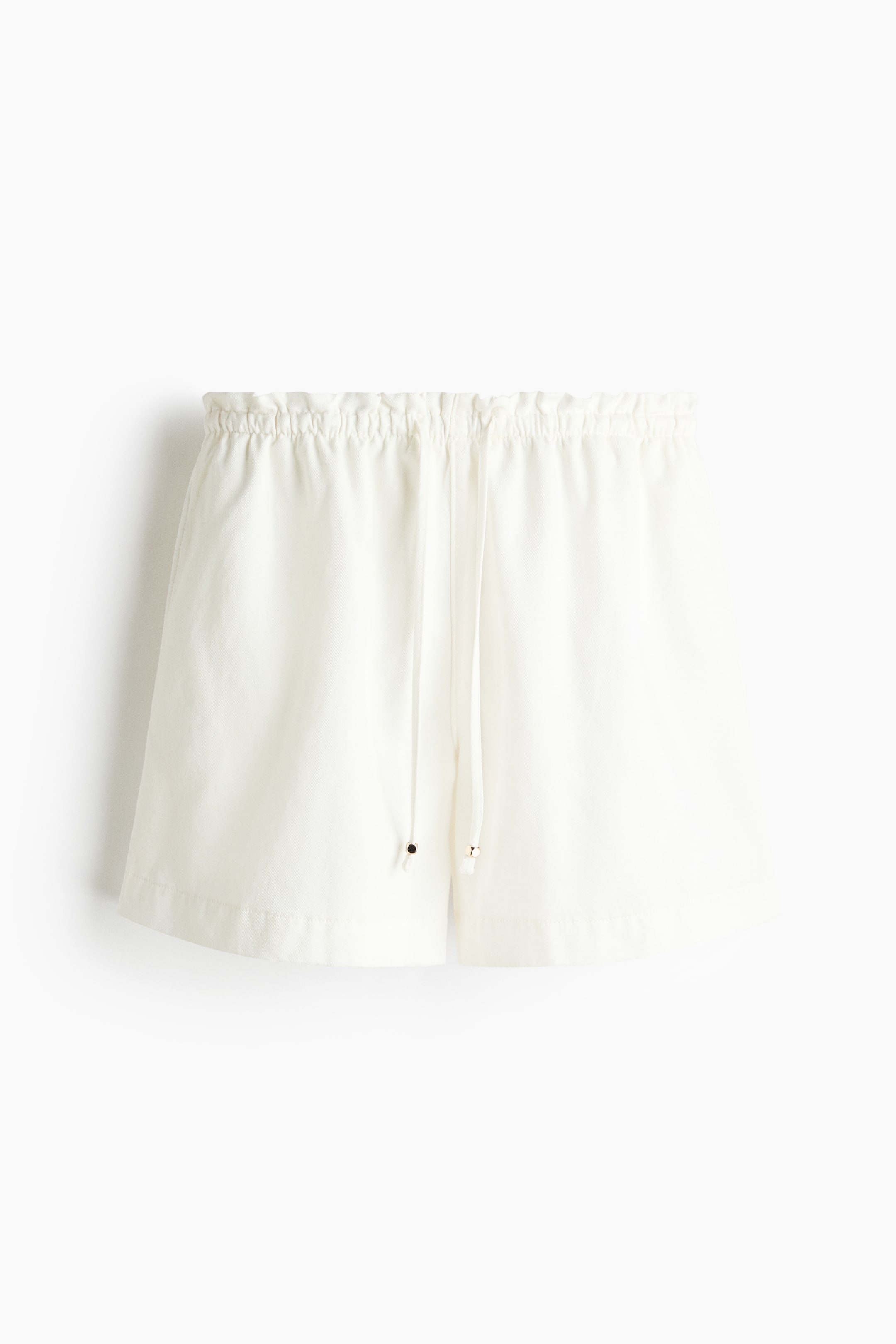 View larger image: Loose-fit white shorts are showcased from the front, featuring a high, elasticated waist with a delicate frill trim and a slender drawstring with metal beads at the centre front. Discreet side pockets are visible along the seams.