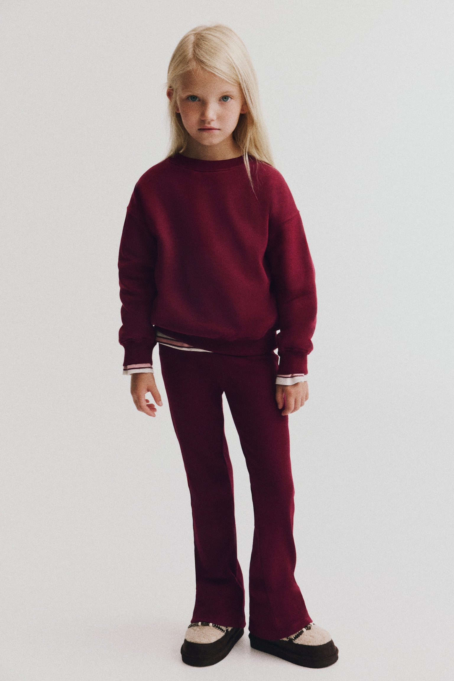 A burgundy two-piece set, shown from the front, features a crewneck sweatshirt and matching ribbed leggings. The sweatshirt has dropped shoulders with a striped shirt layered underneath at the cuffs and hem, while the leggings extend into flared hems. - 1