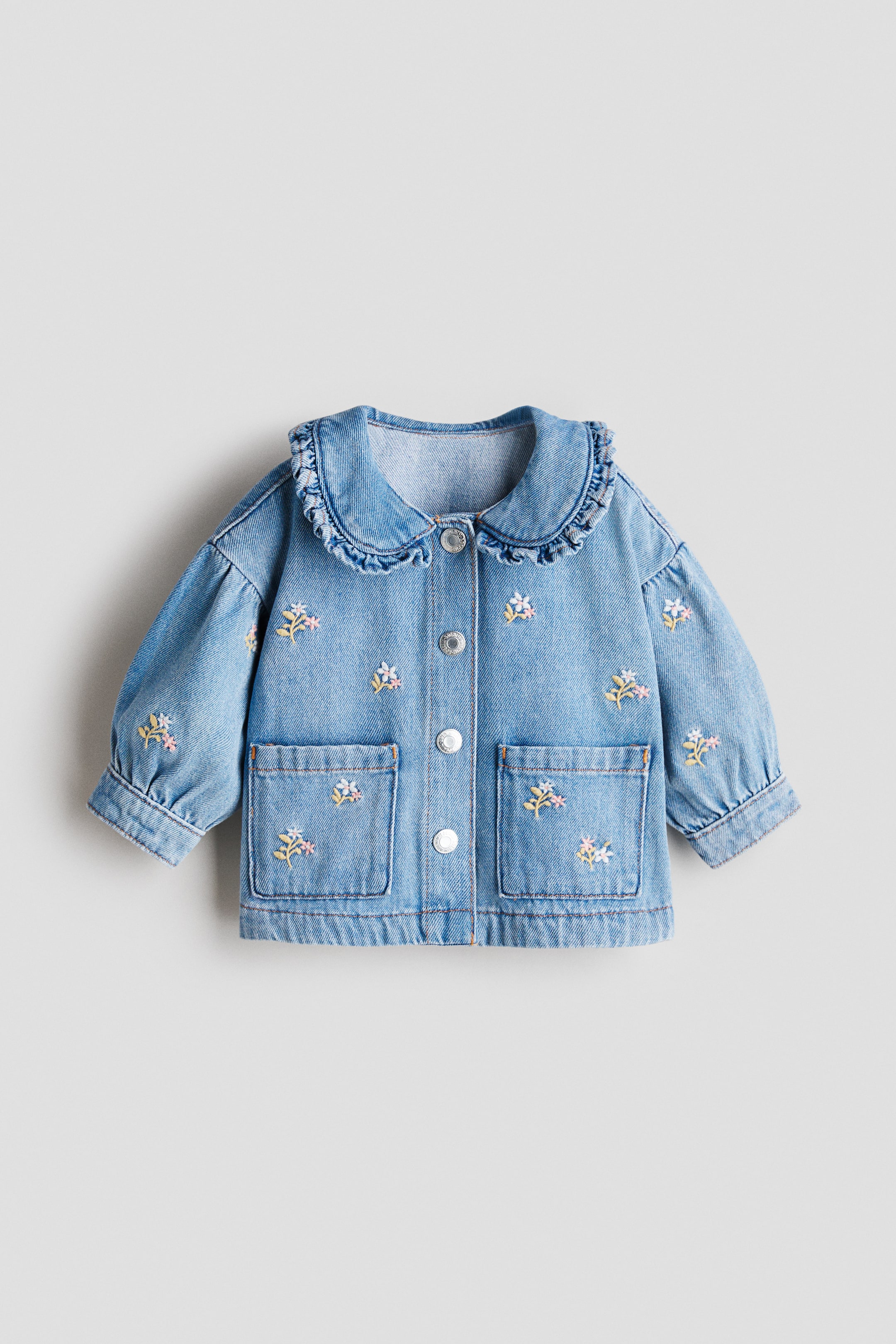 Agrandir l'image: Une veste en denim bleu clair, vue de face, présente un grand col à volants et une fermeture boutonnée sur le devant avec des boutons argentés. Elle comporte deux poches plaquées à l'avant et une broderie florale composée de petites fleurs blanches, roses et jaunes dispersées sur le devant et les manches, ces dernières étant froncées aux poignets.