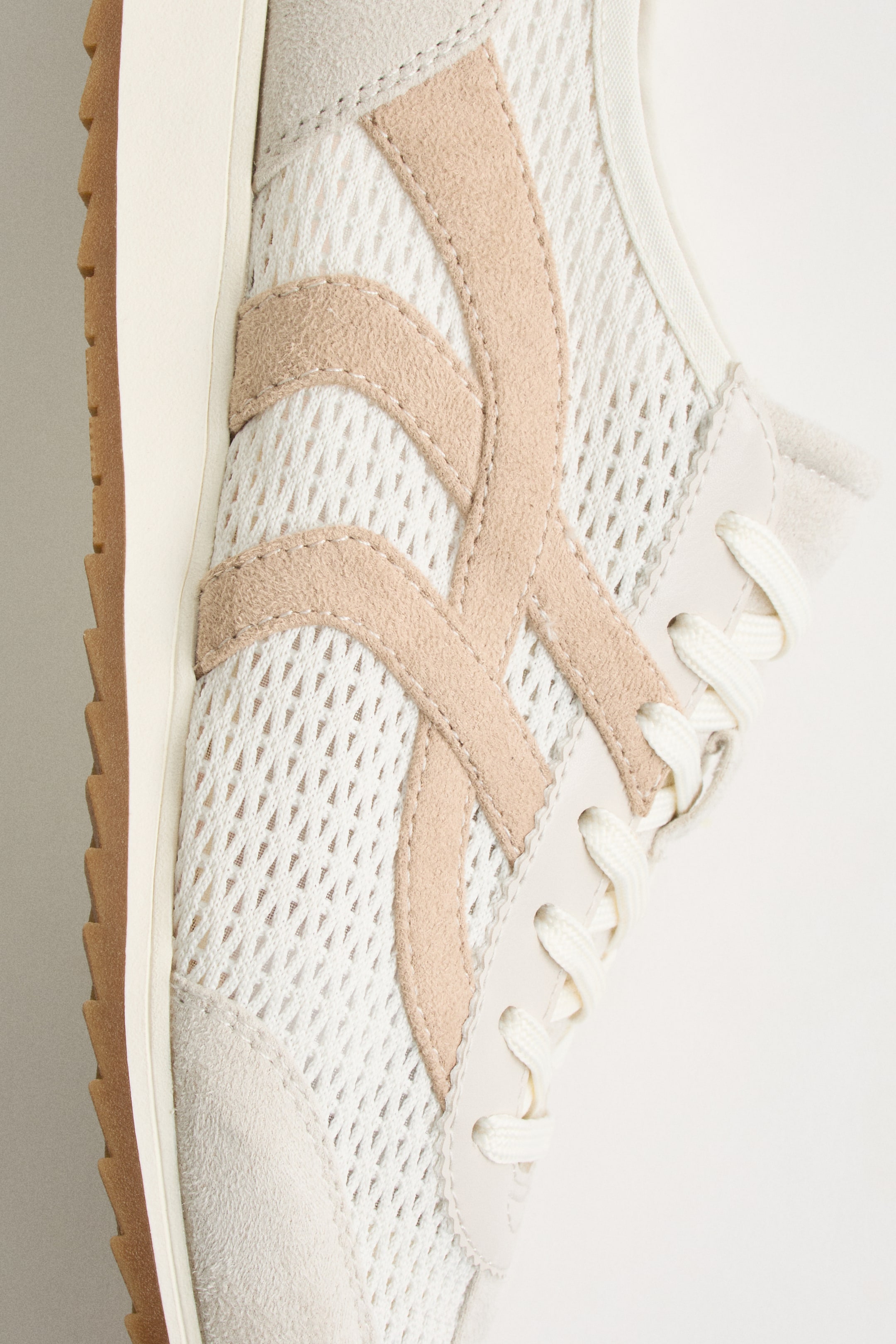 Größeres Bild ansehen: Ein Sneaker präsentiert sein Seitenprofil und zeigt ein weißes Mesh-Obermaterial mit hellbeigen Wildleder-Overlays, die ein geschwungenes Muster über den Mittelfuß, auf der Zehenkappe und um das Schnürsystem bilden. Cremefarbene Schnürsenkel sind durch Ösen gefädelt, und eine cremefarbene Zwischensohlenkante ist über einer geriffelten Außensohle sichtbar.