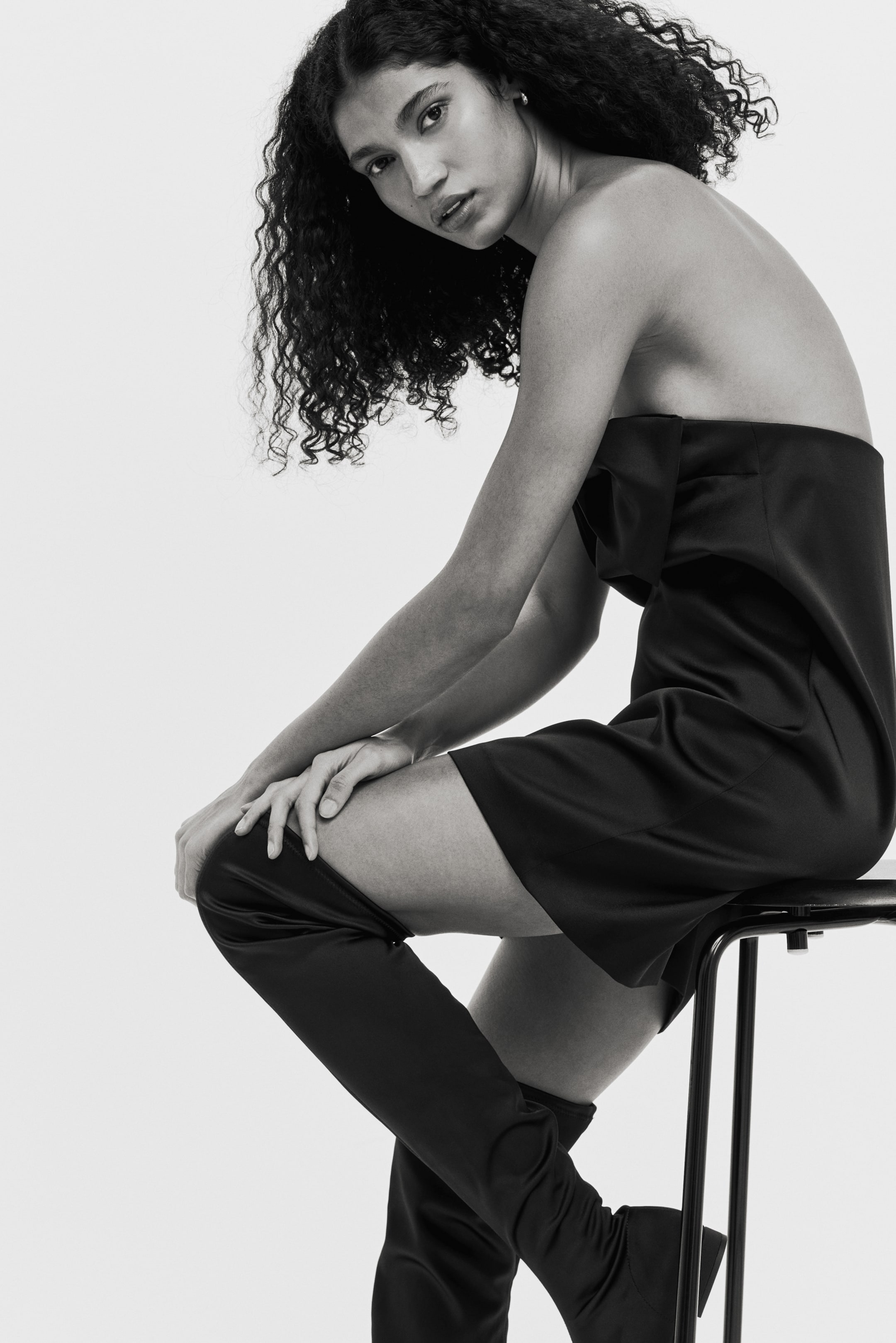 Agrandir l'image: Une robe noire sans bretelles présente un détail structuré et plissé au niveau du buste et s'étend jusqu'à mi-cuisse, tandis que des bottes noires montantes et lisses épousent étroitement les jambes.
